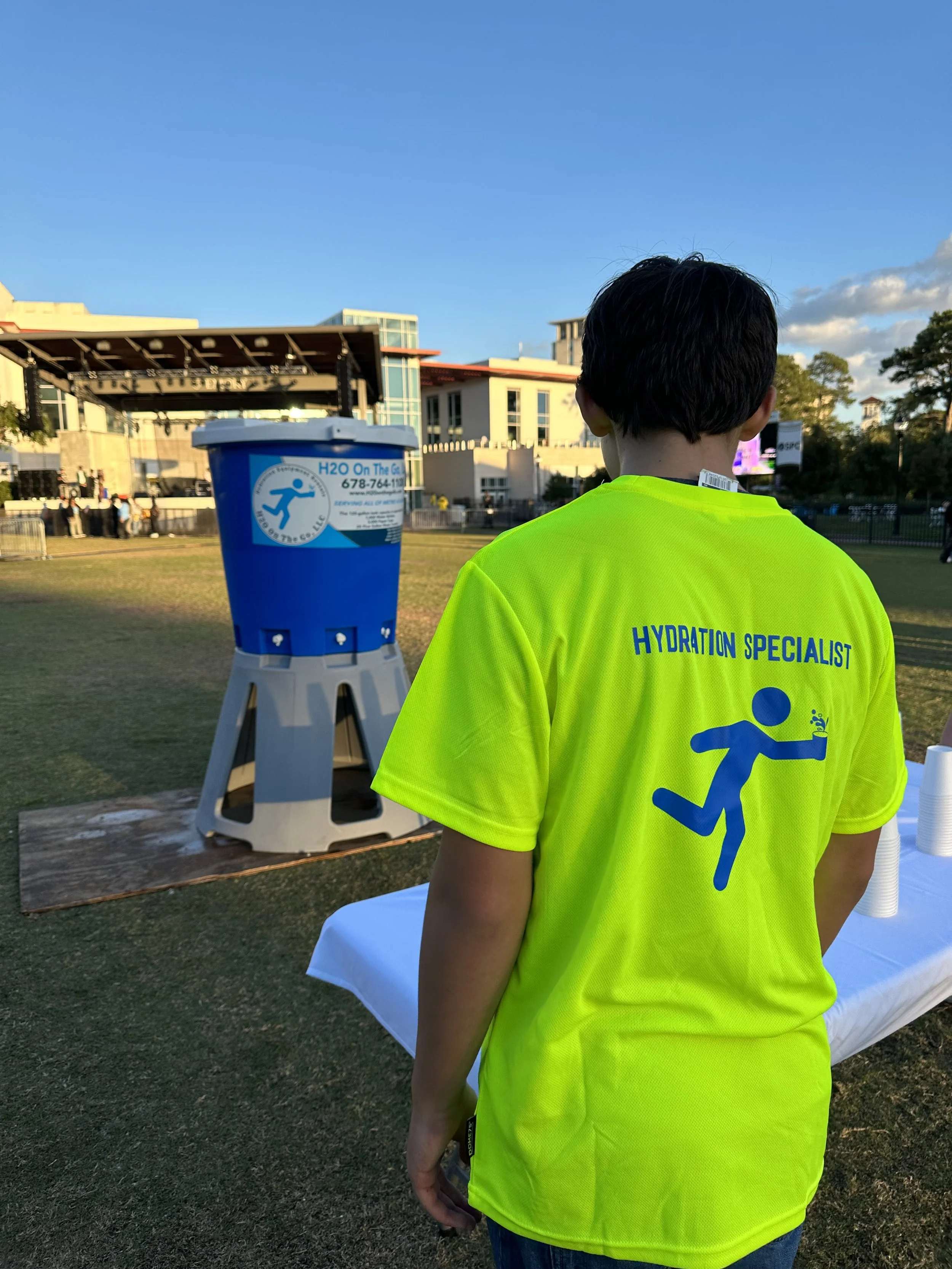 Person wearing a neon yellow shirt with the words 'Hydration Specialist' on the back, standing outdoors near a water station, with a stage and buildings in the background.