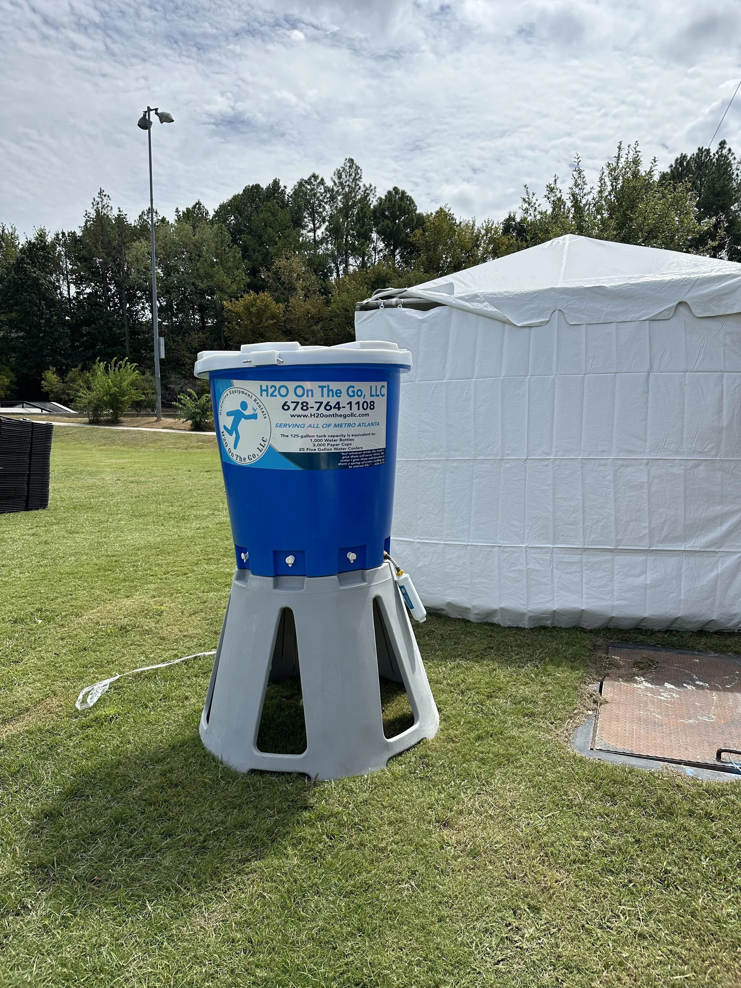 A portable water dispenser with a blue container on a gray stand, labeled "H2O On The Go, LLC," situated on a grassy outdoor area near a white tent and a patch of pavement, with trees and a cloudy sky in the background.