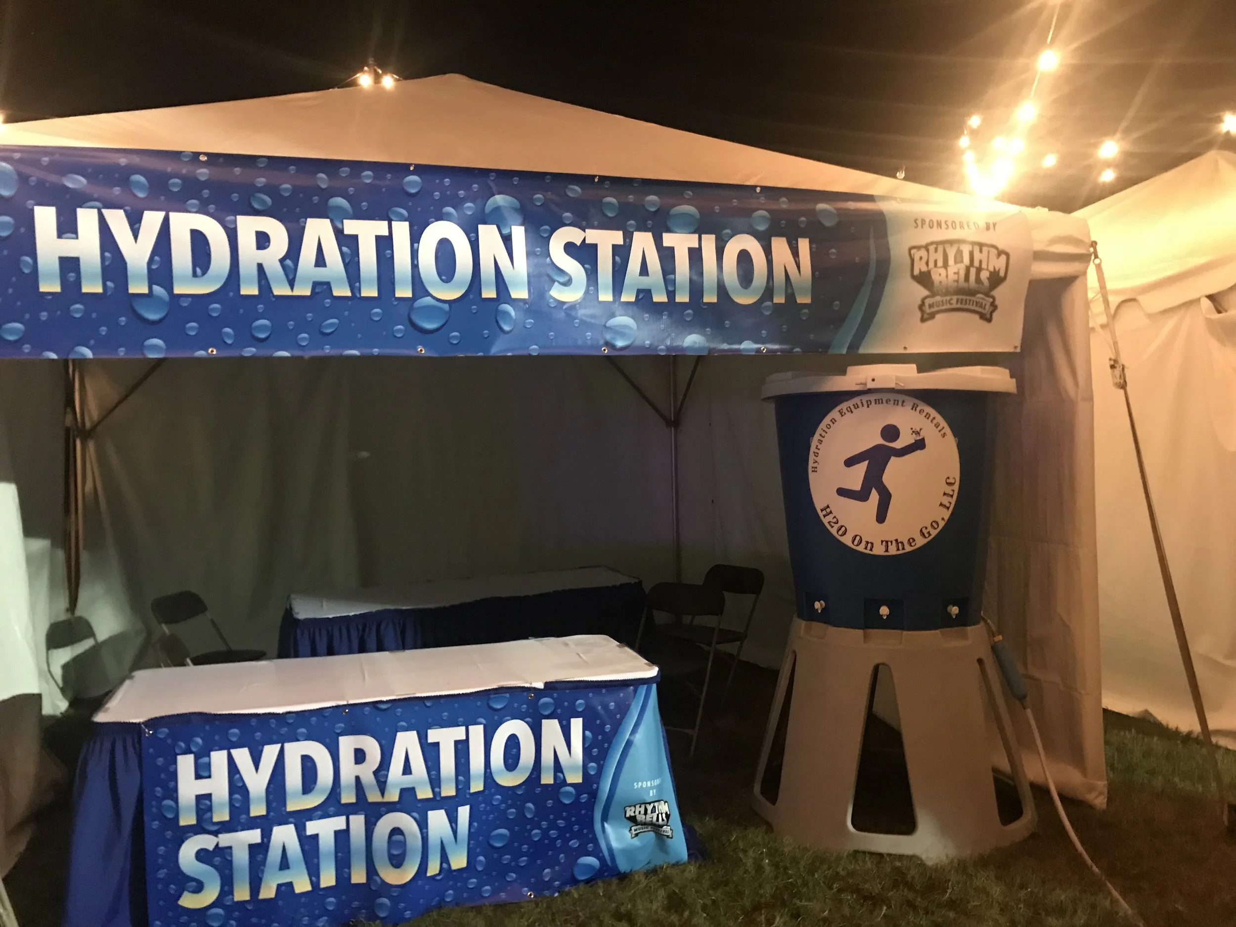 A hydration station at an outdoor event with a large banner reading 'Hydration Station', a table covered in a matching tablecloth, and a large water cooler with a sign that says 'Hydration Equipment Rentals H2O On The GO LLC' next to it.