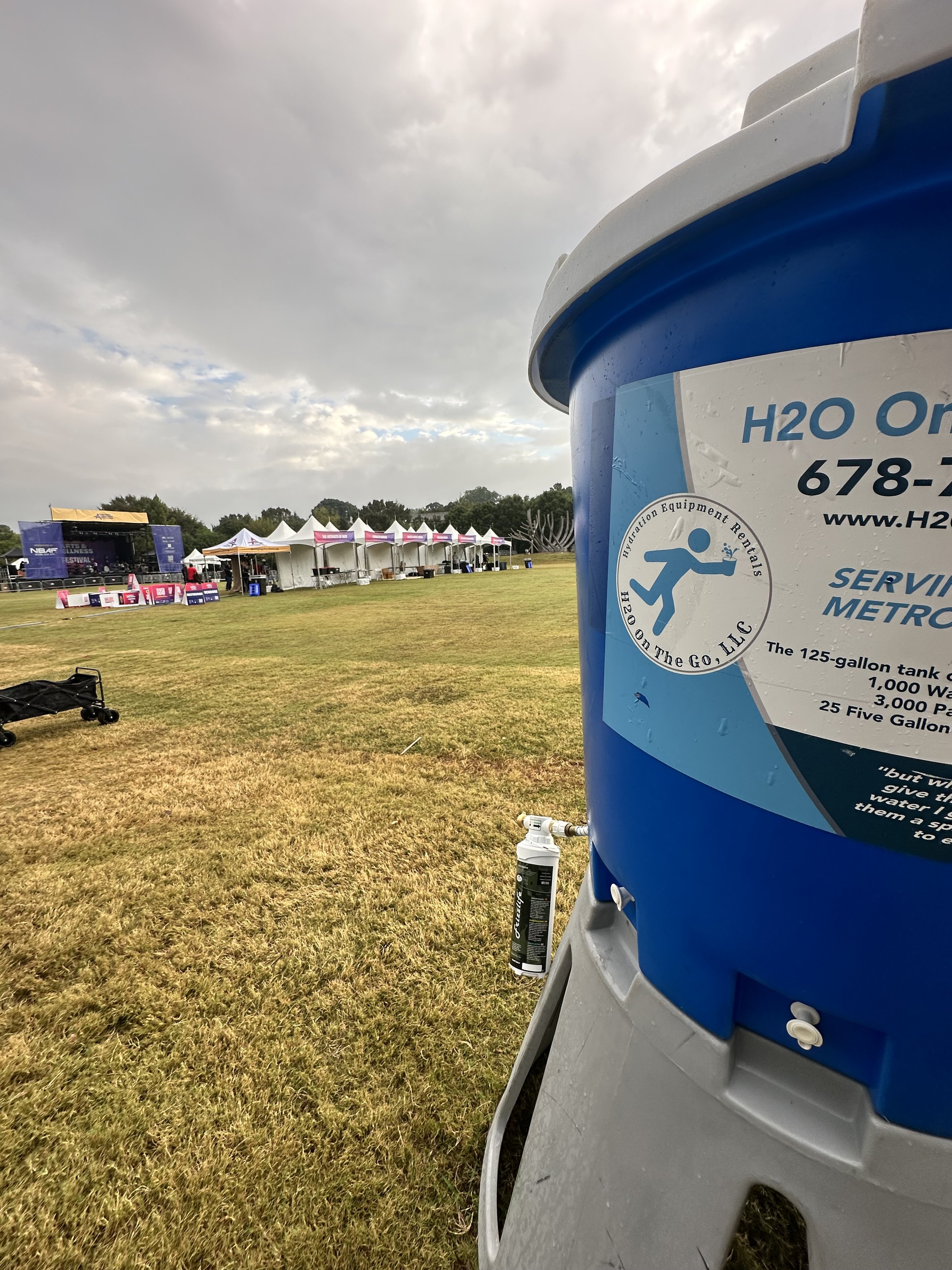 Close-up of a large blue water cooler or dispenser with a company logo and text, set up outdoors on a grassy field, with event tents and a stage in the background under a cloudy sky.