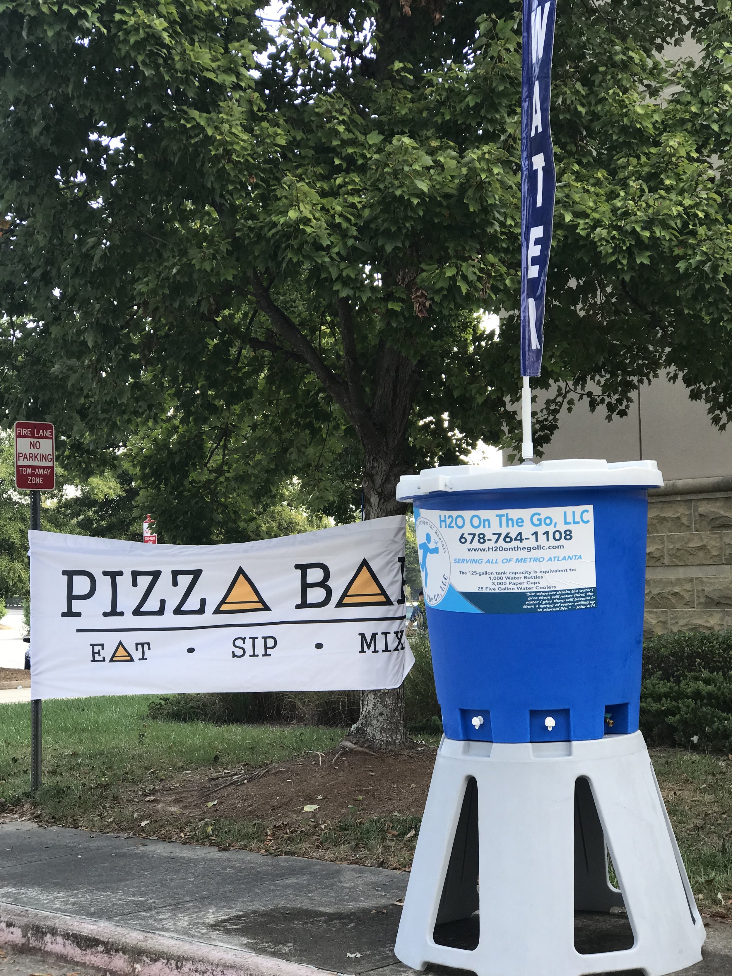 A water vending station with a blue water dispenser and a flag, located next to a tree and a banner promoting pizza and beer.