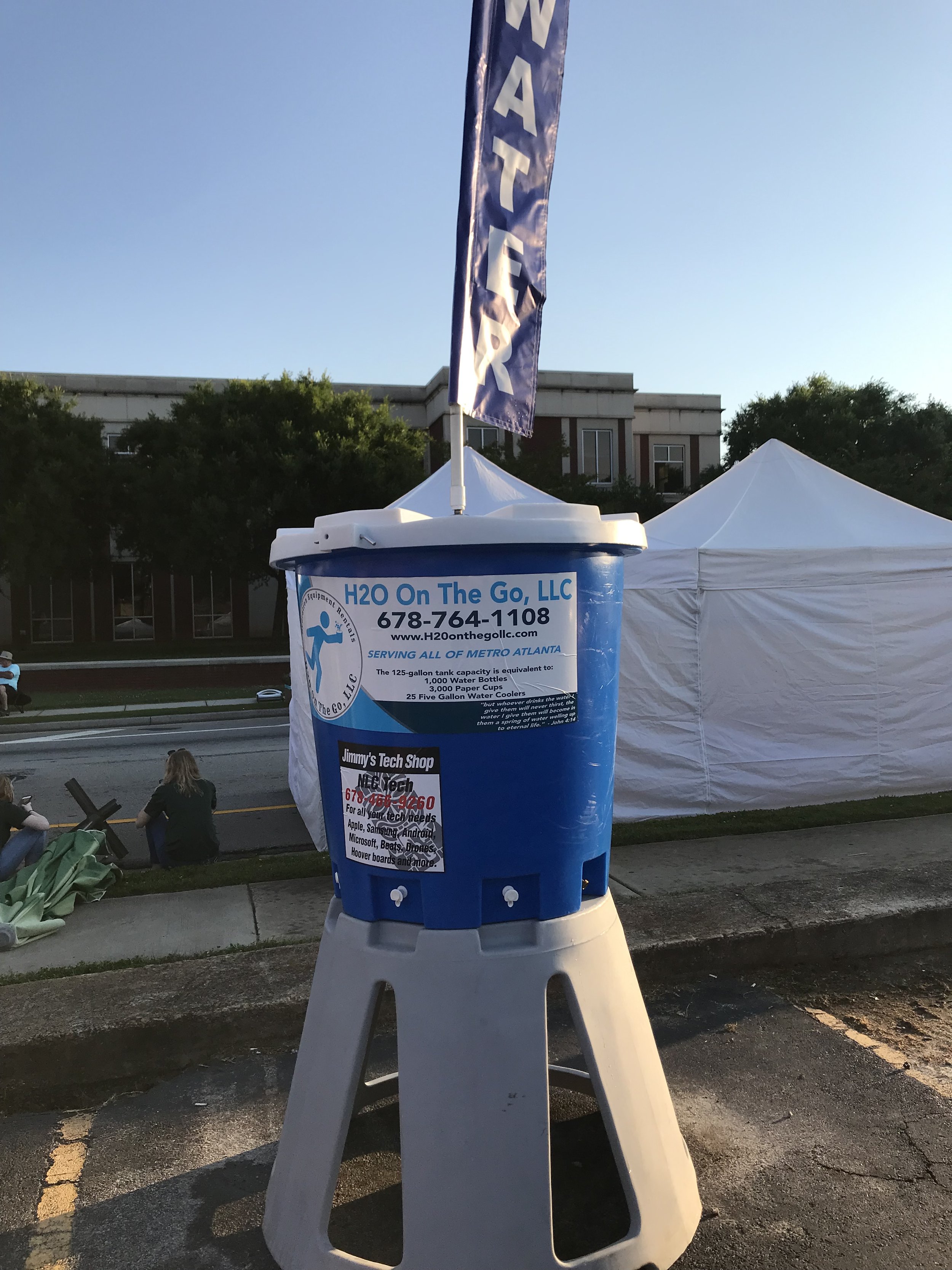 Blue water cooler with signs and banner for 'H20 On The Go, LLC,' serving metro Atlanta, with a tent and people sitting in the background.