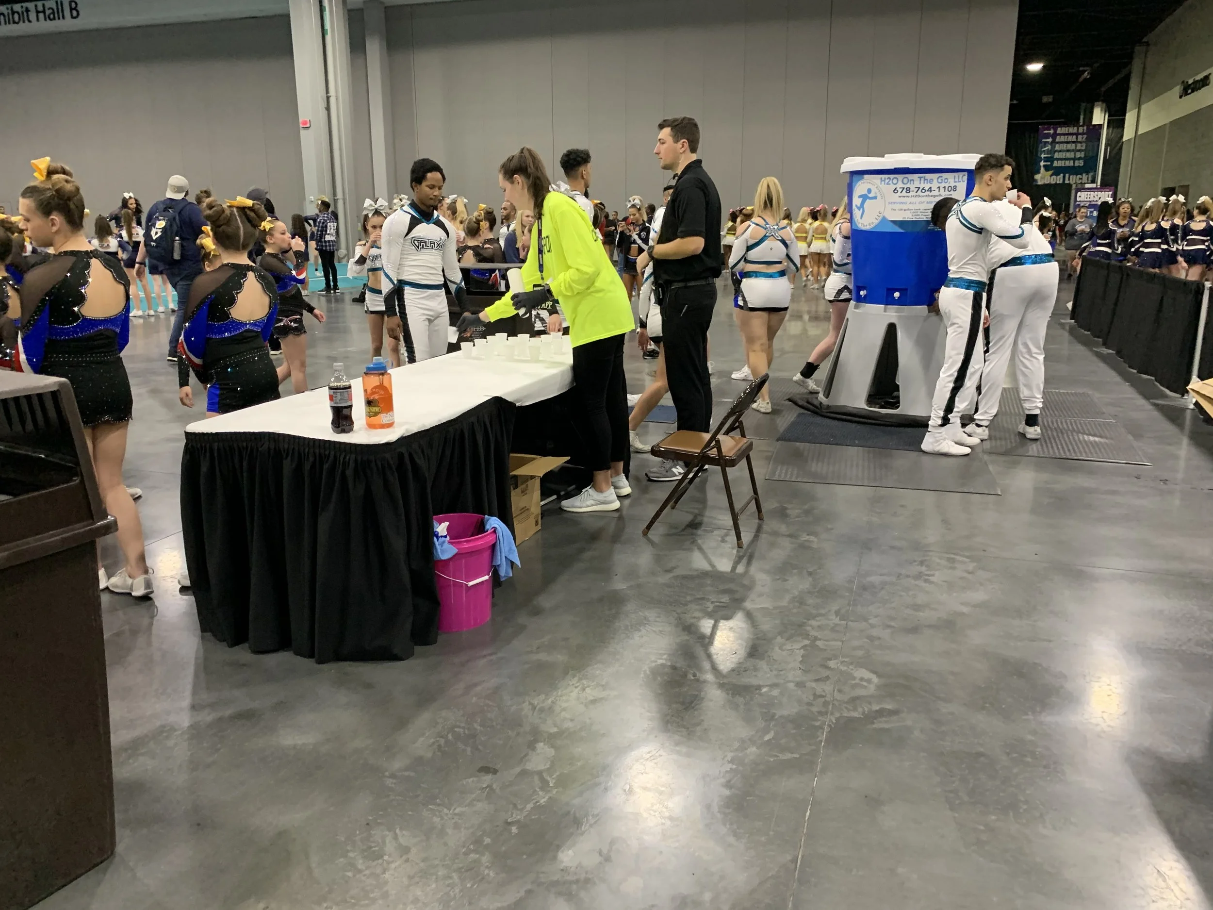 Cheerleaders and athletes in a large indoor sports arena, with a hydration station and groups in uniforms preparing for a cheerleading event.