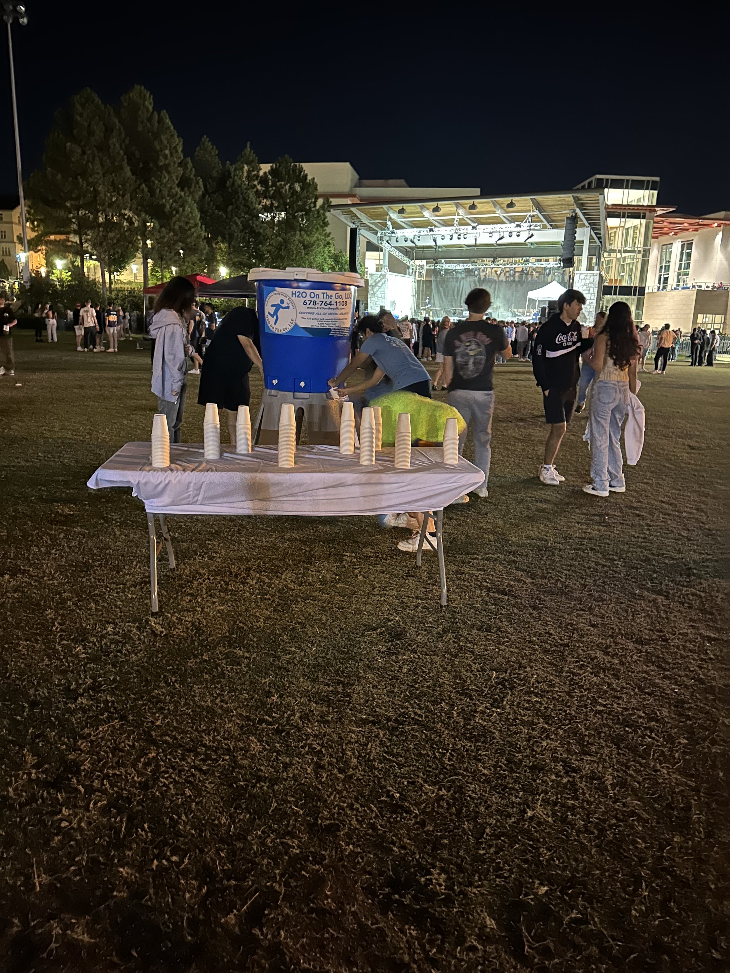 Night scene of a public outdoor event with a stage in the background. In the foreground, a table with stacked paper cups and a water dispenser, with people nearby. The area is lit, with trees and a crowd of people in front of the stage.
