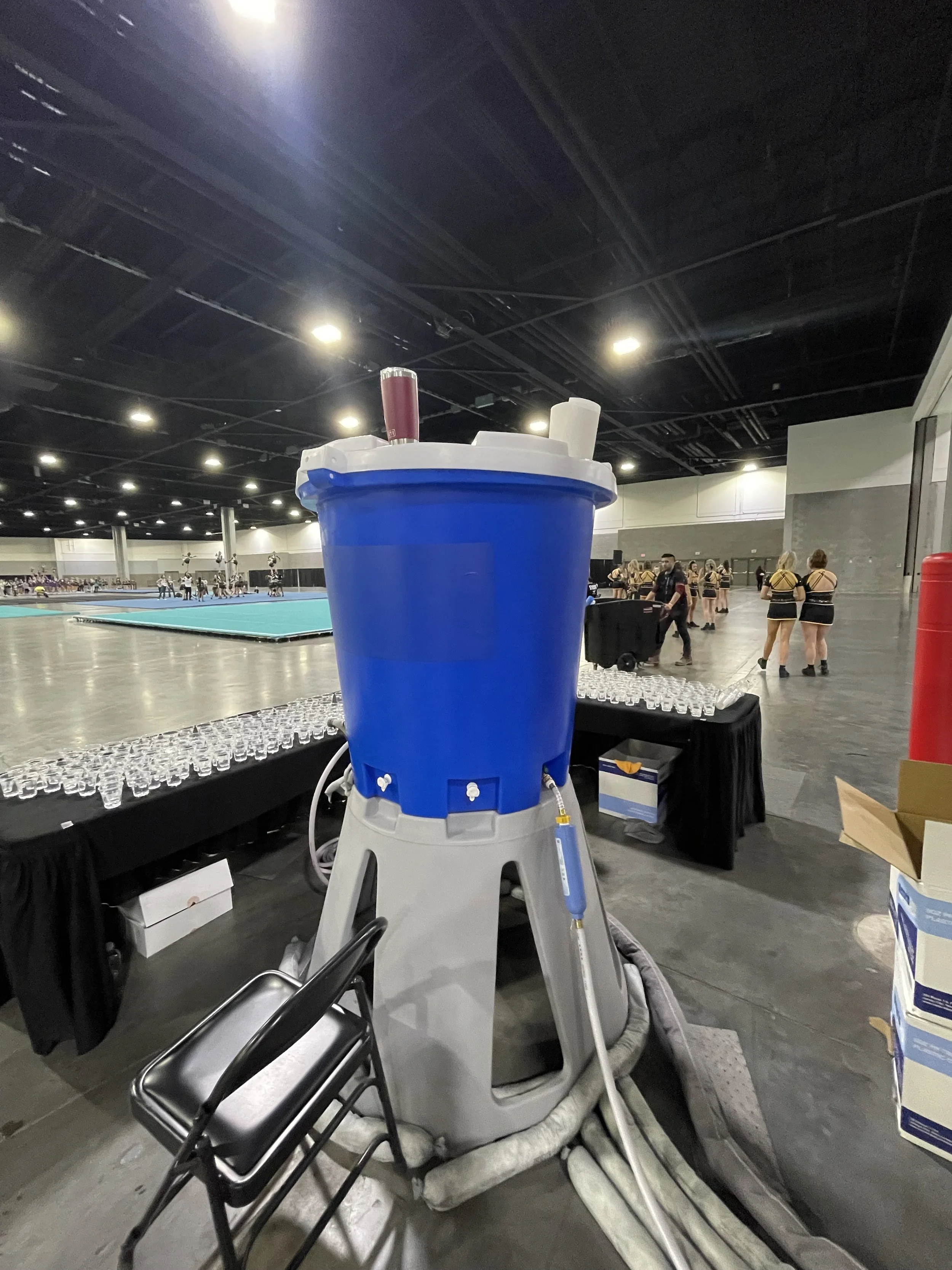 A large blue water cooler with a gray base and white and maroon cups, situated in an indoor sports venue with a cheerleading squad practicing in the background.