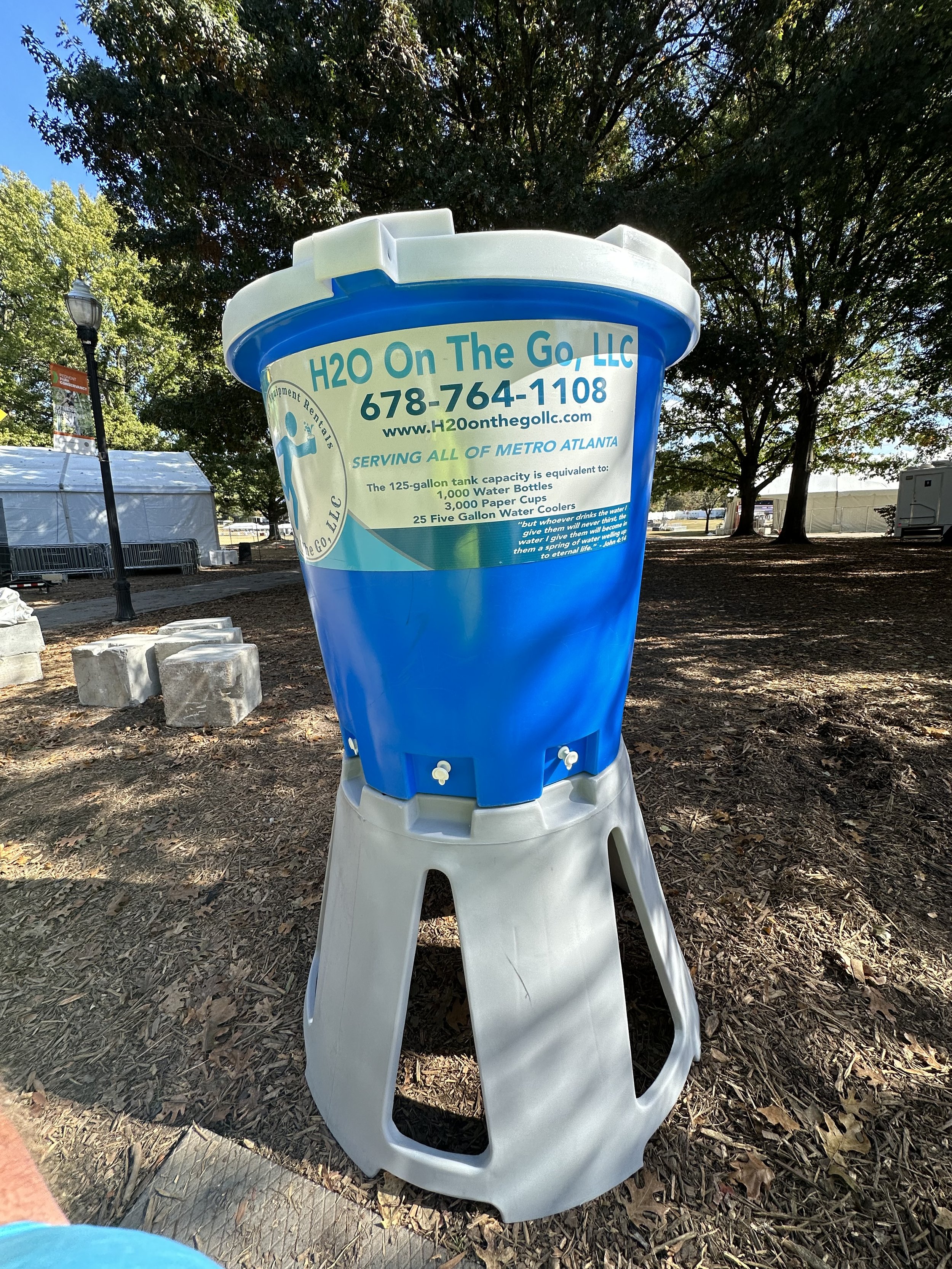 A large advertisement fountain for 'H2O On The Go, LLC' standing outdoors with trees in the background. It is designed to look like a big water cup with informational text about water delivery services.