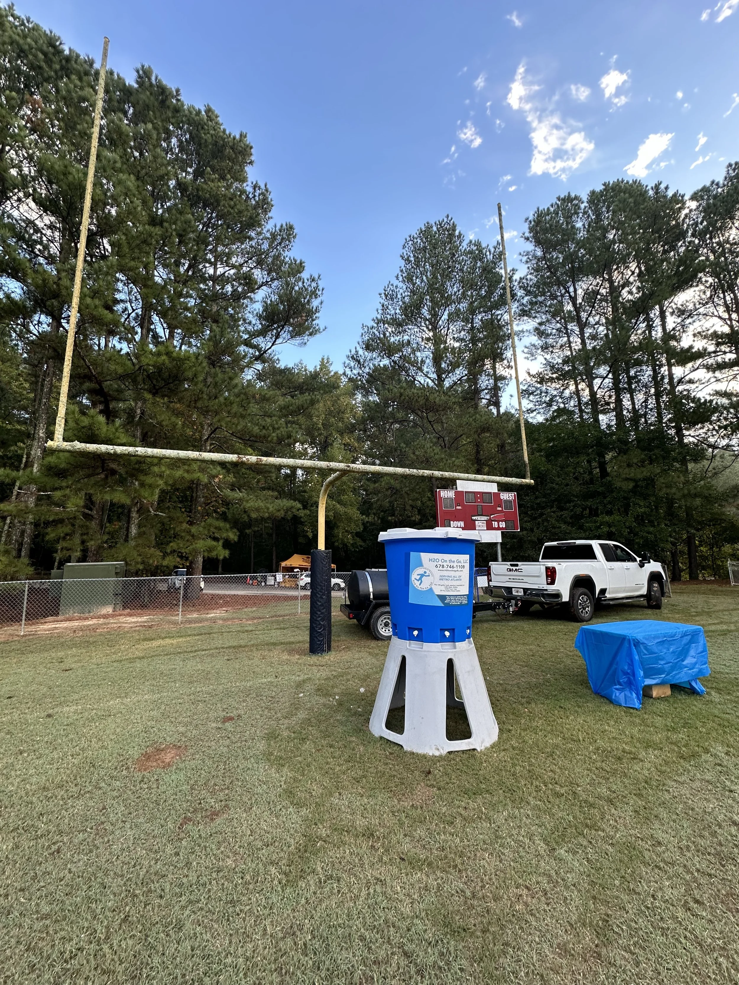A football field with a goalpost, a blue water station, a white pickup truck, and a scoreboard in the background, surrounded by trees.