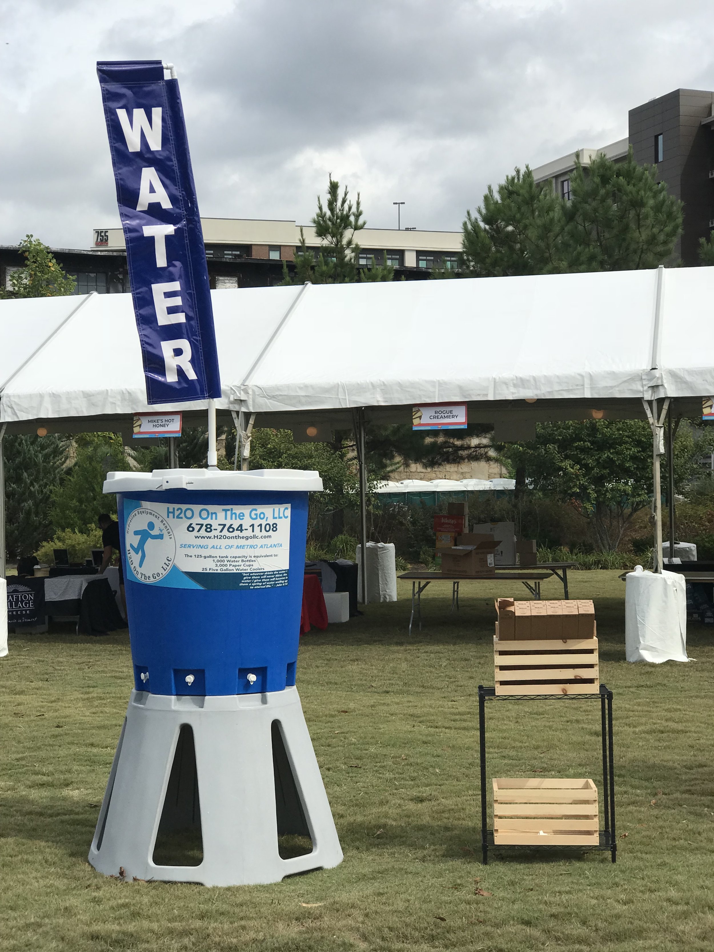 A large water dispenser with a blue sign that reads 'H2O On The Go, LLC' and phone number, set outdoors on a grassy area in front of a white tent. The tent has signs labeled 'Mike's Hot Honey' and 'Rogue Creamery', with several tables and boxes under