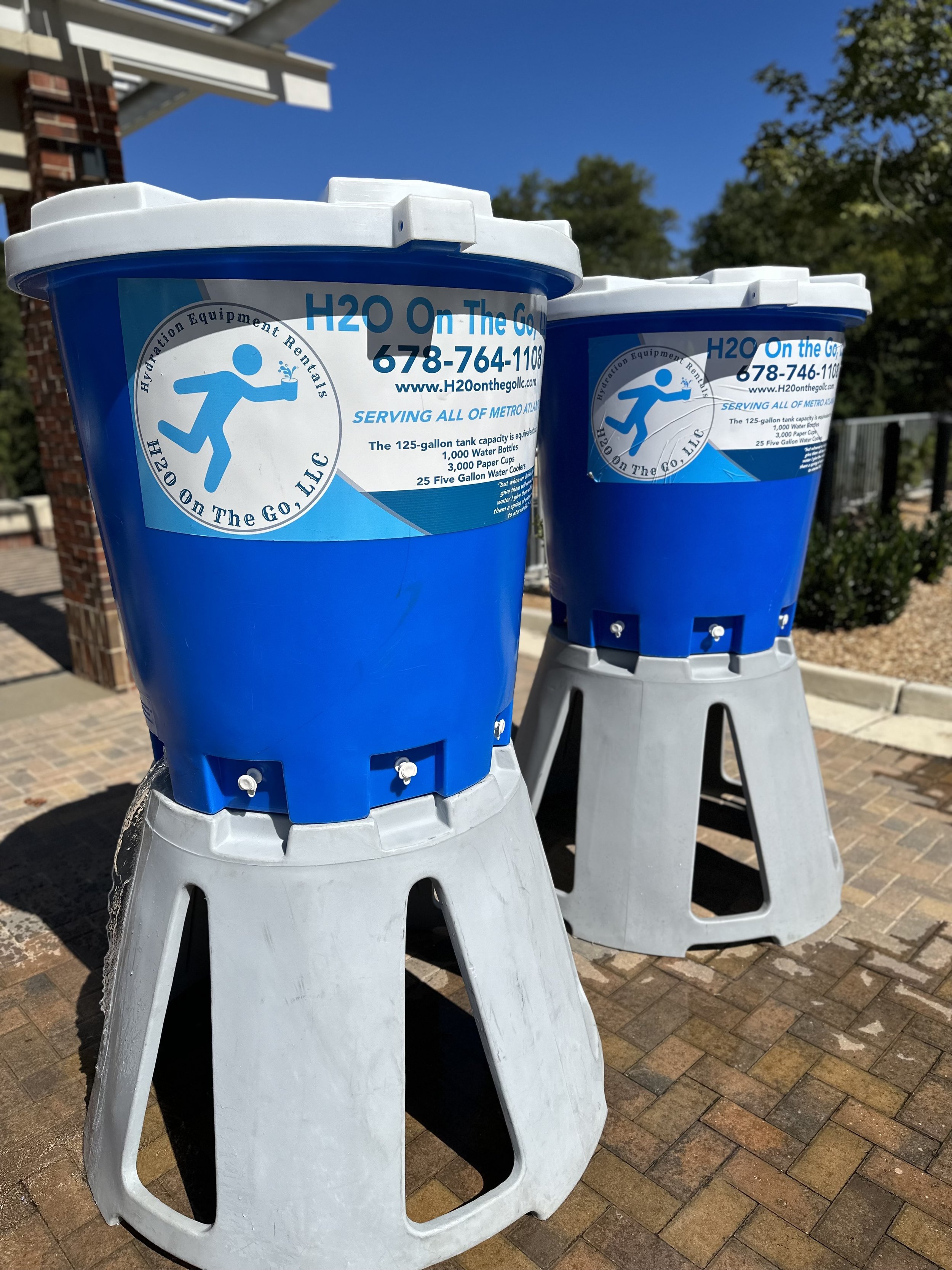 Two large blue water refill stations on gray stands outside under a clear blue sky, with a building, trees, and a brick sidewalk in the background.