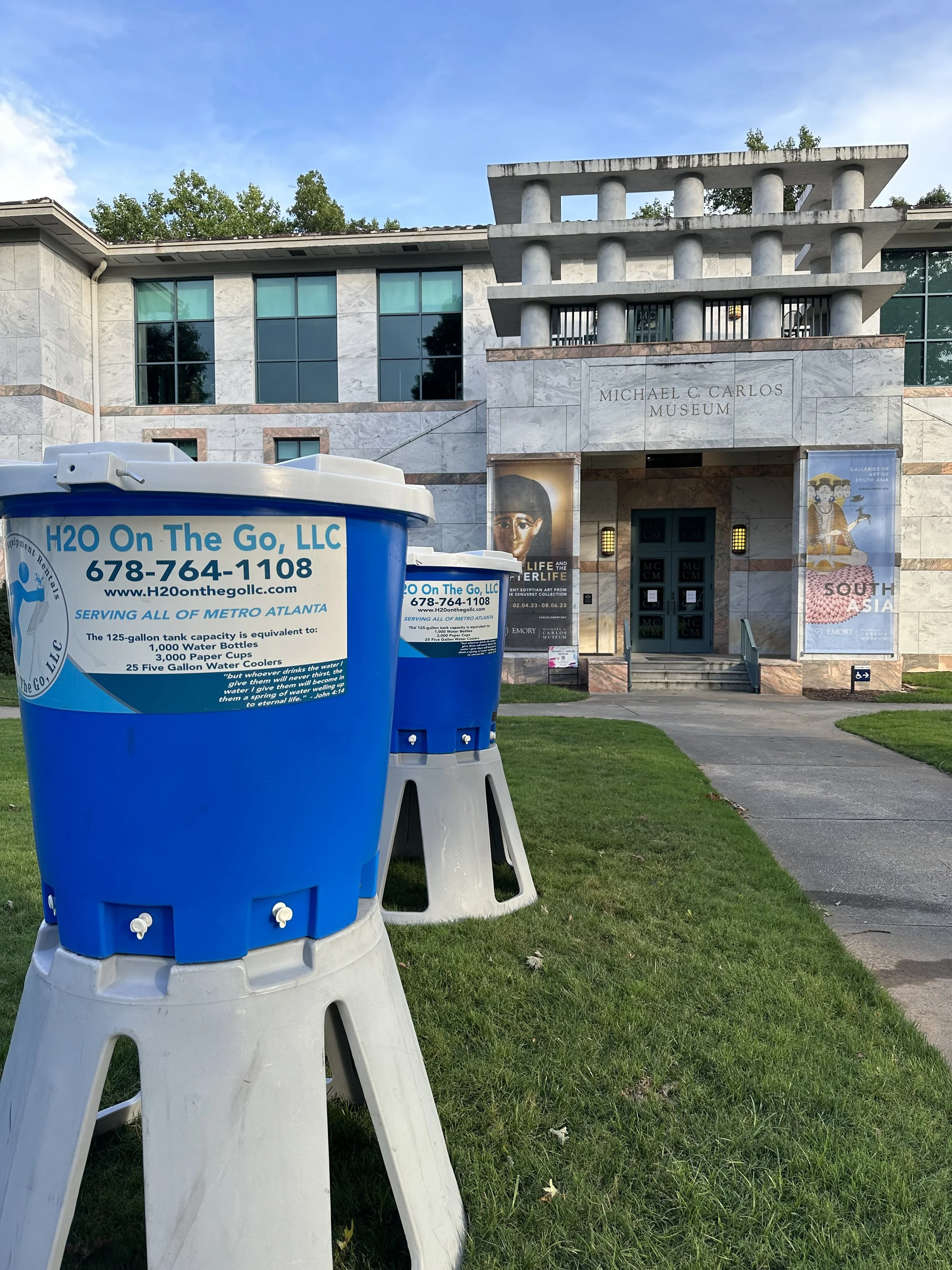 Two blue water cooler jugs outside the Michael C. Carlos Museum, with the museum building in the background and a walkway leading to the entrance.