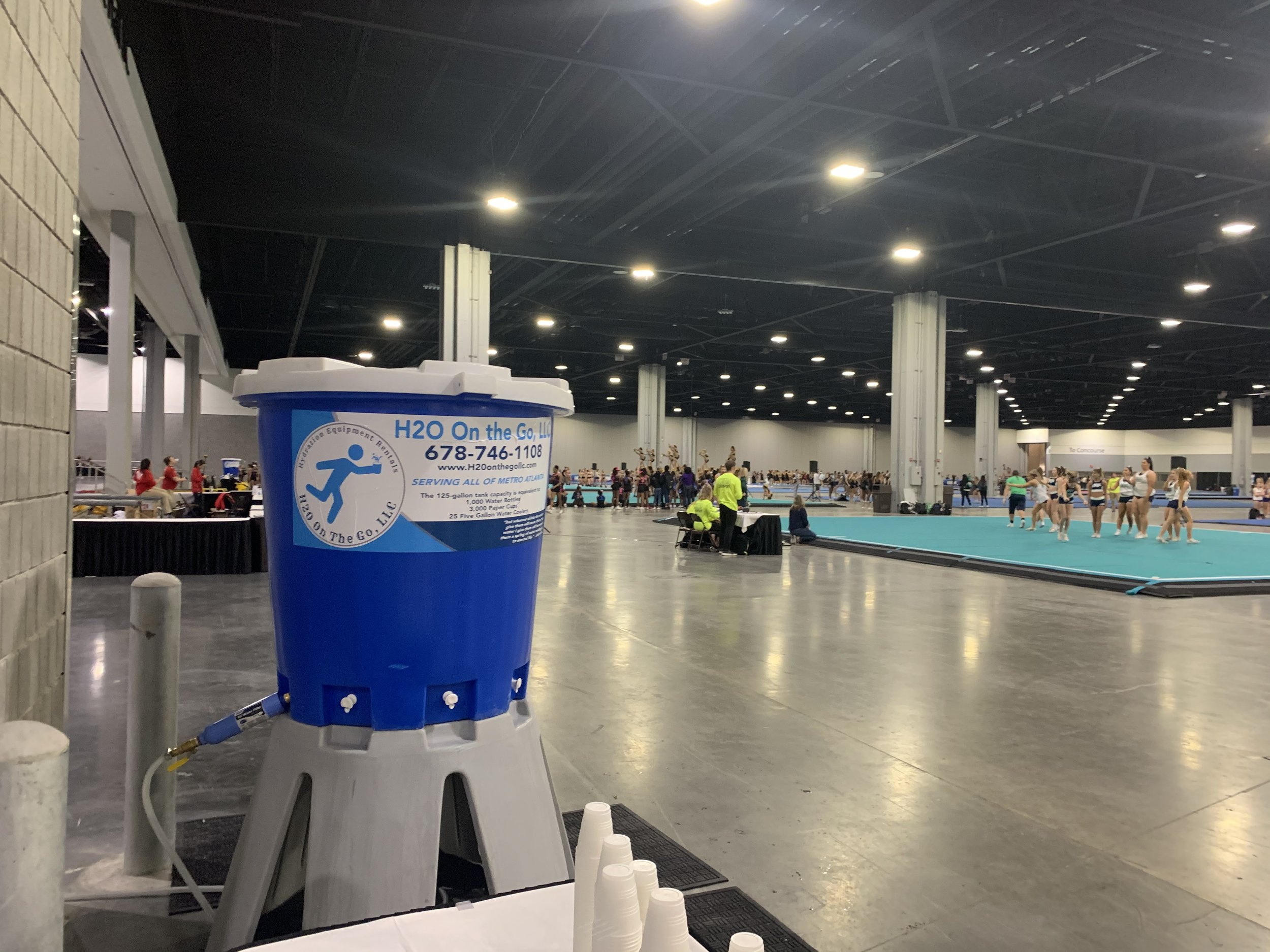 A large indoor gymnastics event with many athletes warming up or competing on mats. In the foreground, a large blue water cooler with a company logo and contact information. To the left, there is a table with cups, and in the background, spectators a
