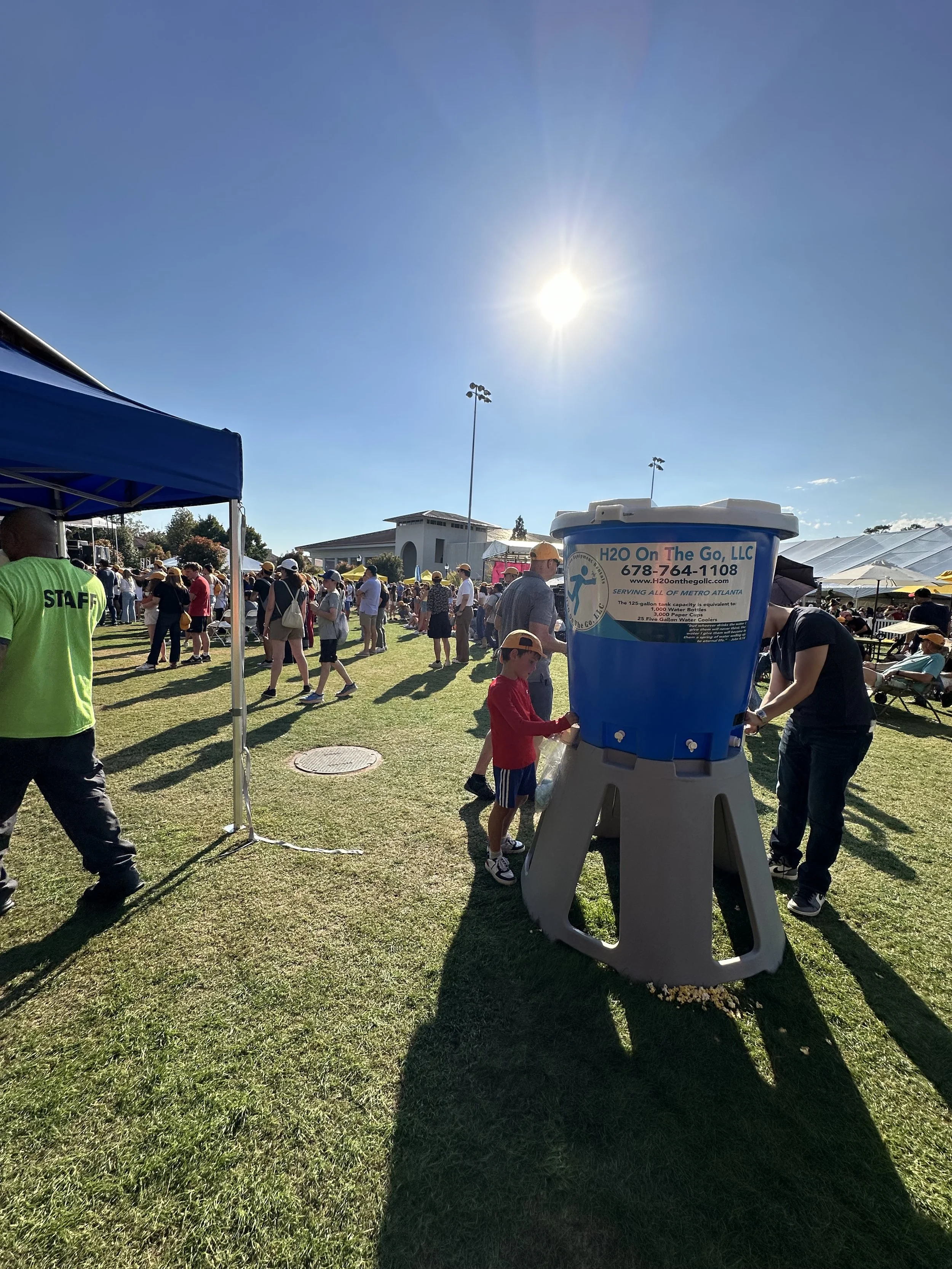 A water refill station at an outdoor event with many attendees, including children and adults, under a clear sunny sky.