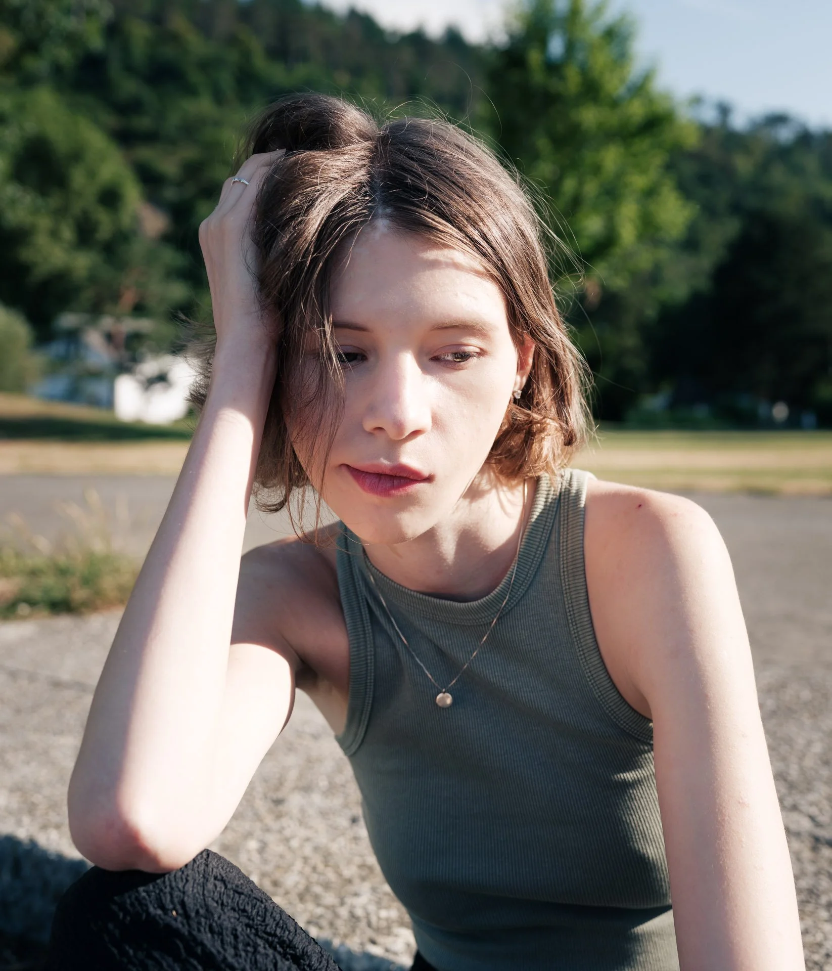 Close-up of a woman with light skin and short brown hair, wearing a grey sleeveless top and a silver necklace, sitting outdoors on a sunny day with green trees and a park in the background.