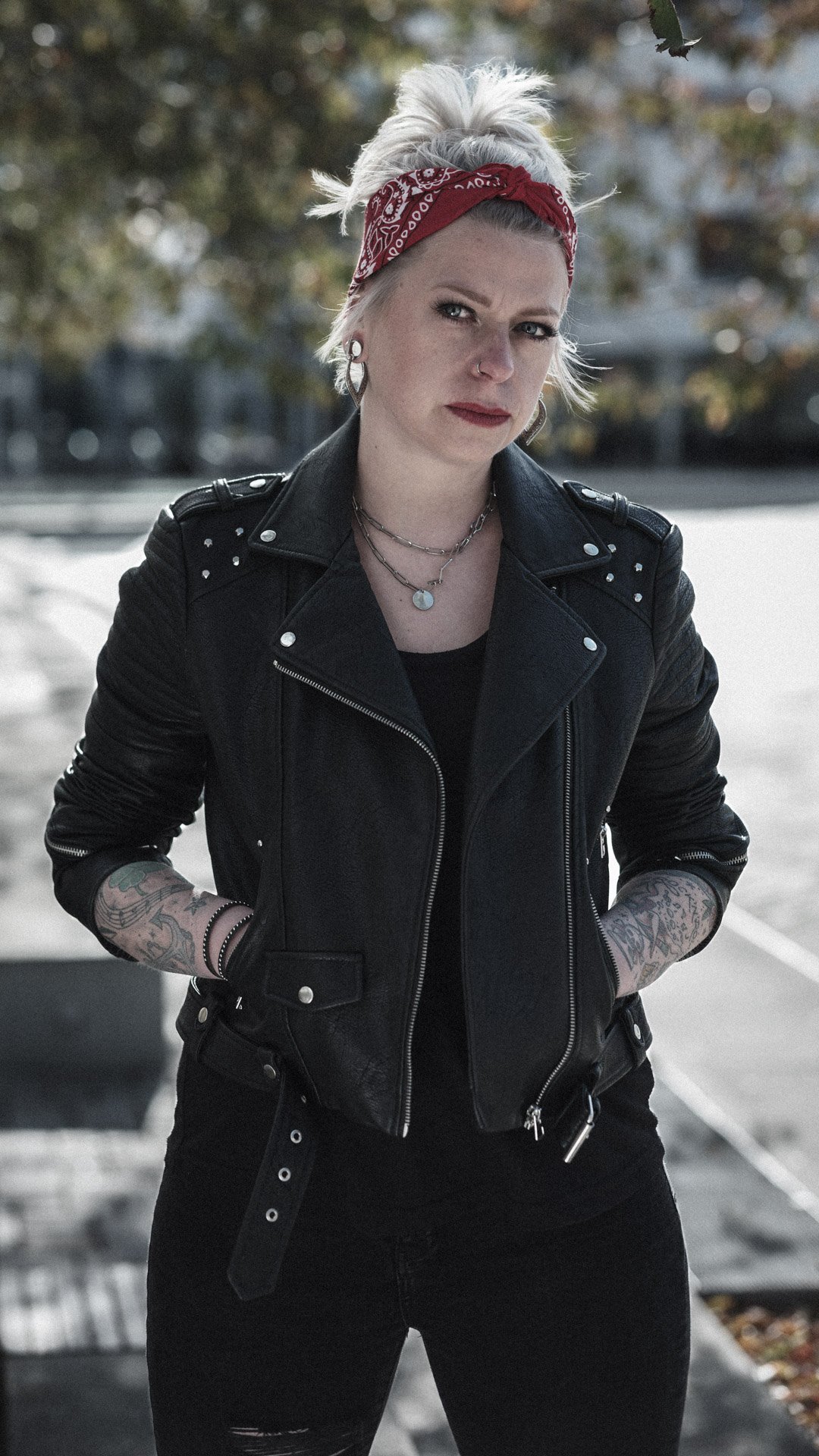 A blonde woman wearing a red bandana, black leather jacket with silver studs, and black jeans, standing outdoors with trees in the background.