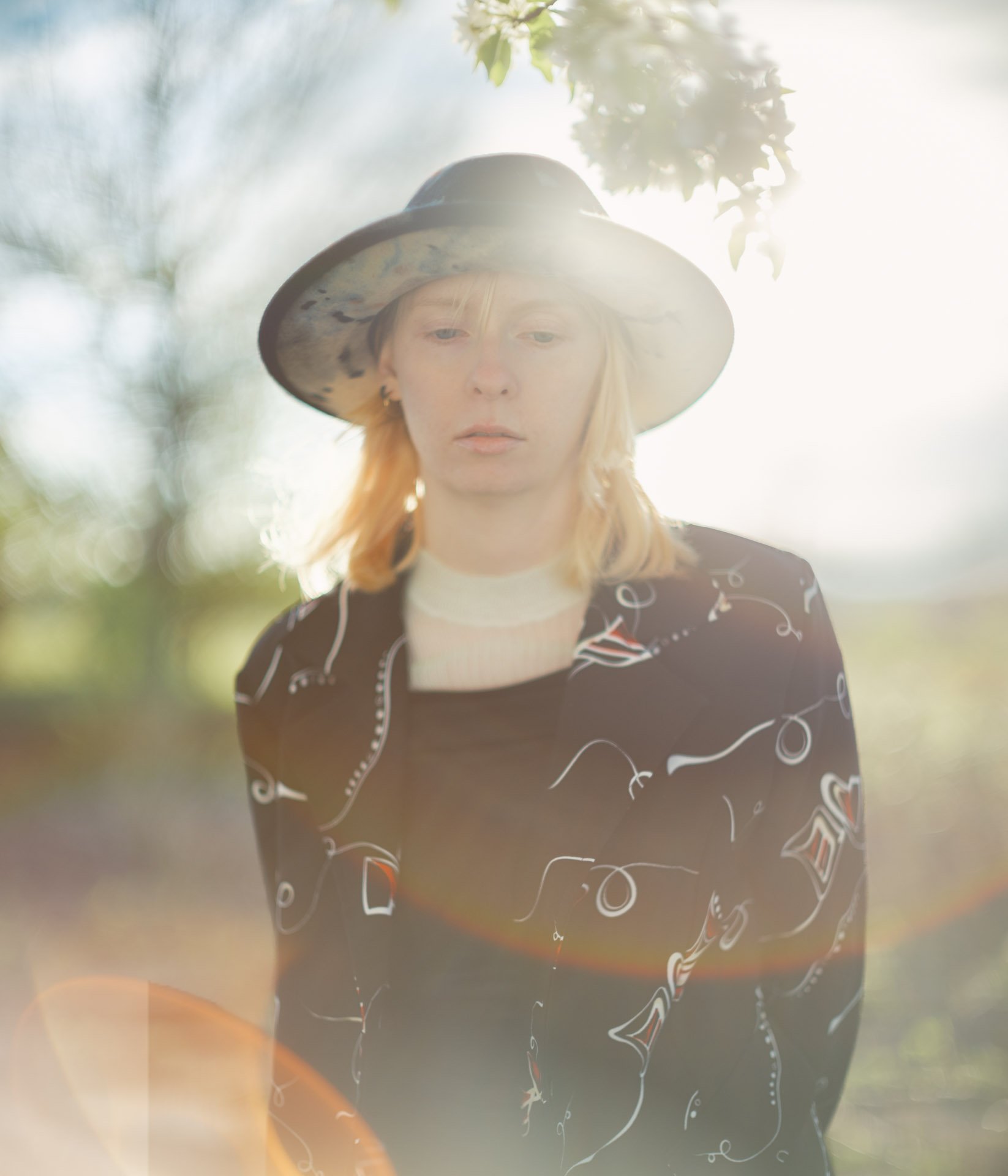 A woman with blonde hair wearing a wide-brimmed hat and a black dress with white and red abstract patterns, standing outdoors with trees and sunlight in the background.