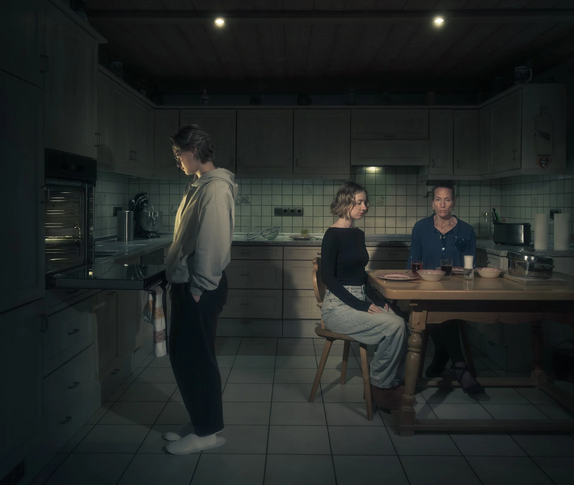 Three women in a kitchen, one standing near a stove and two sitting at a table, with various kitchen appliances and bowls on the table.