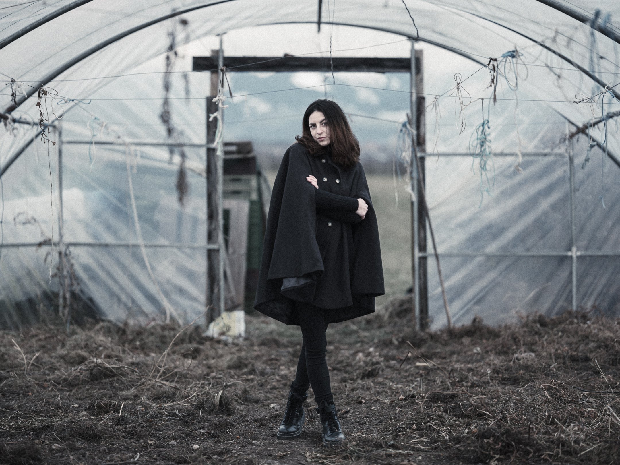 A woman standing with her arms crossed inside a greenhouse structure with a cloudy sky in the background.