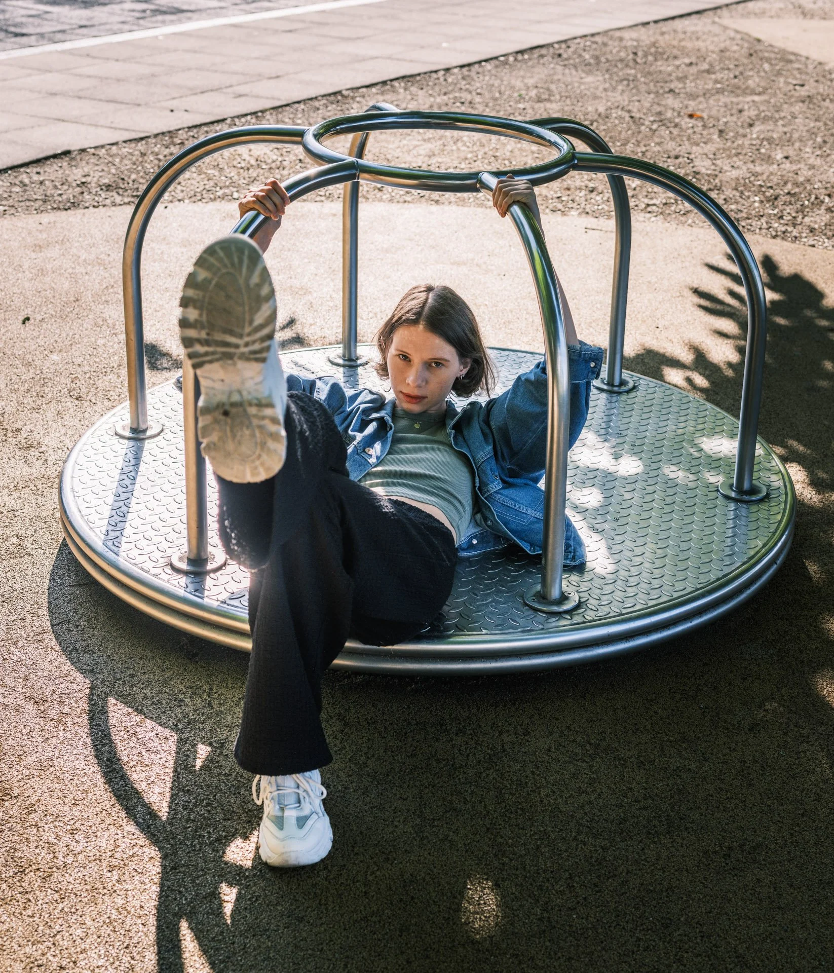 A young girl in casual clothes and white sneakers lying on a metal playground merry-go-round, holding onto the bars with a determined expression.