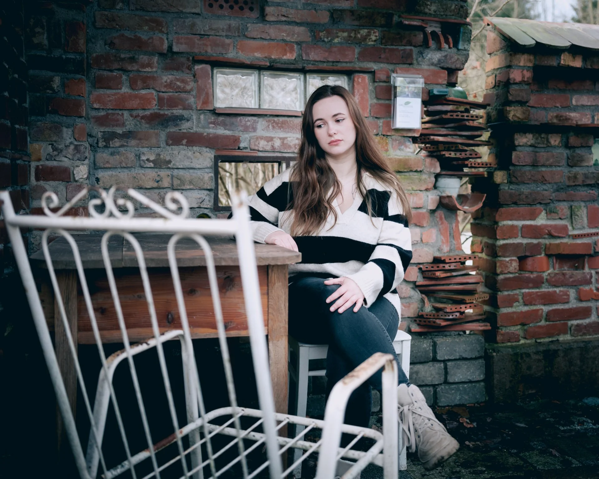 A young woman with long brown hair, wearing a black and white striped sweater, sits on a white chair outdoors against a brick wall, with a distressed white chair and a wooden table in front of her.