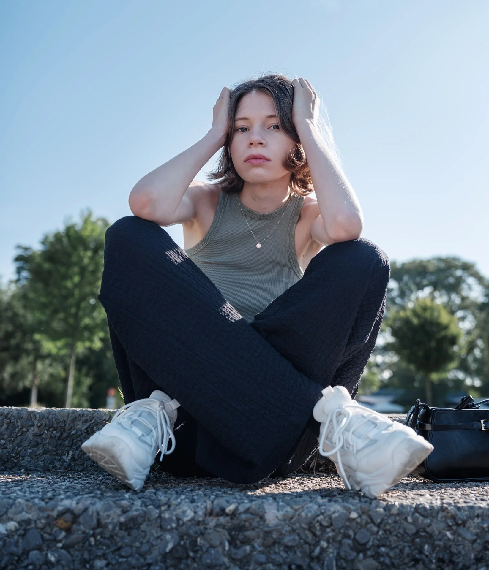 Young woman sitting on a concrete surface outdoors with her hands on her head looking at the camera, wearing a sleeveless top, dark pants, and white sneakers, with trees and a clear sky in the background.