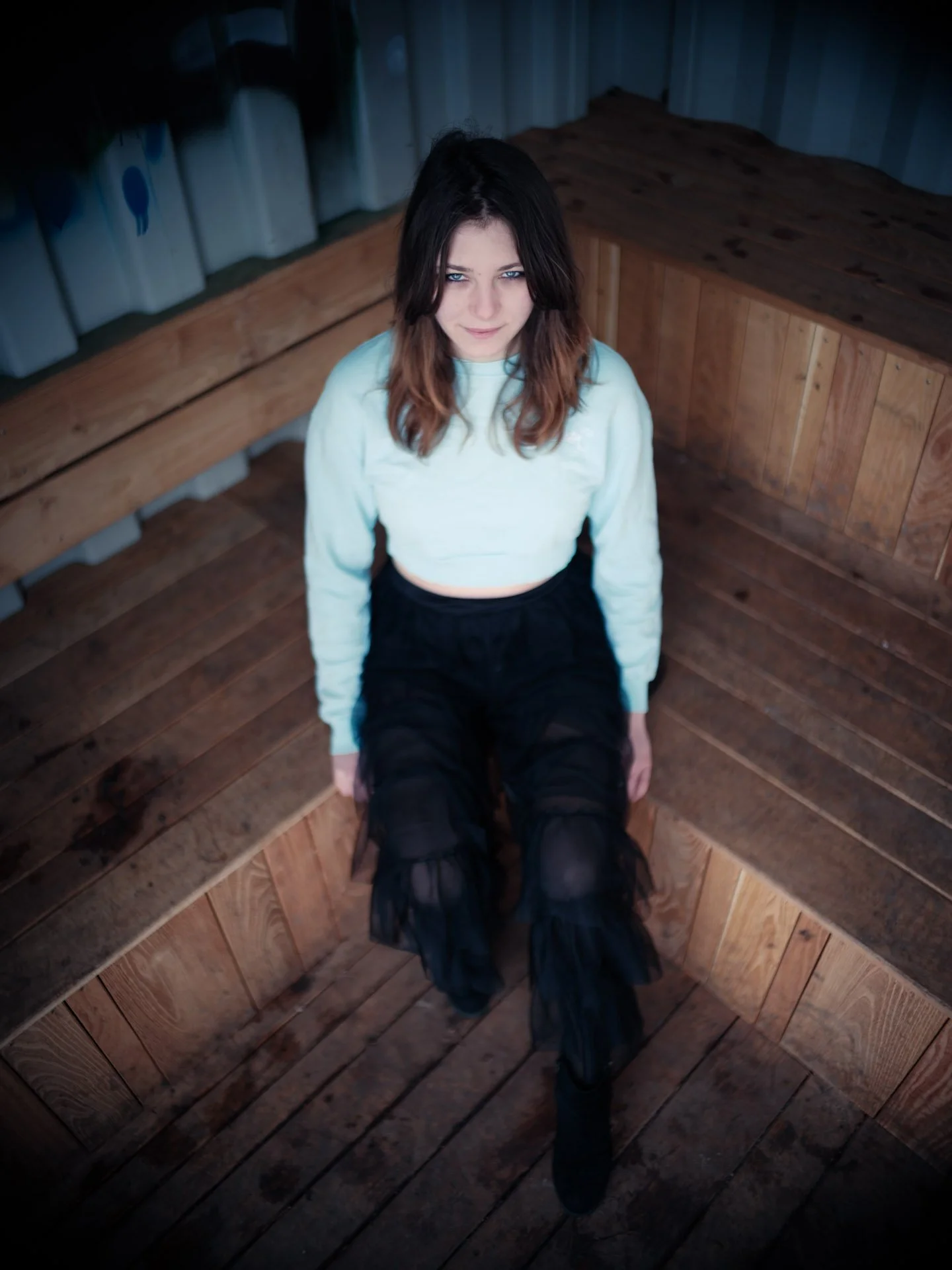 A young woman with shoulder-length dark hair and blue eyes sits on a wooden circular bench, looking up at the camera with a slight smile. She is wearing a light-colored sweatshirt and black pants. The background features a curved wooden wall.