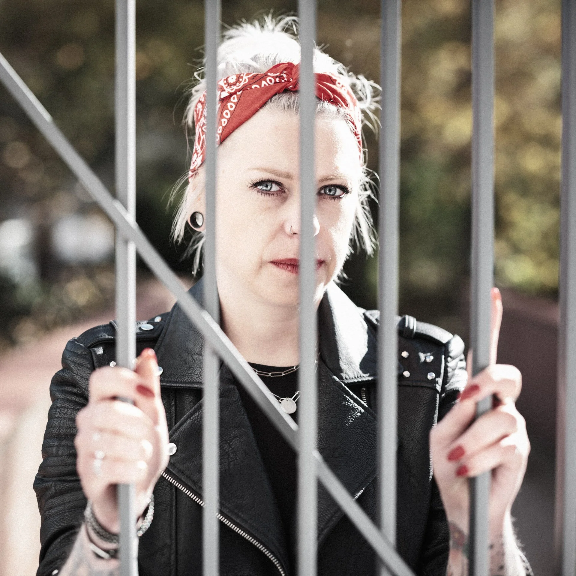 A woman with platinum blonde hair, red bandana, and leather jacket looking through a metal fence.