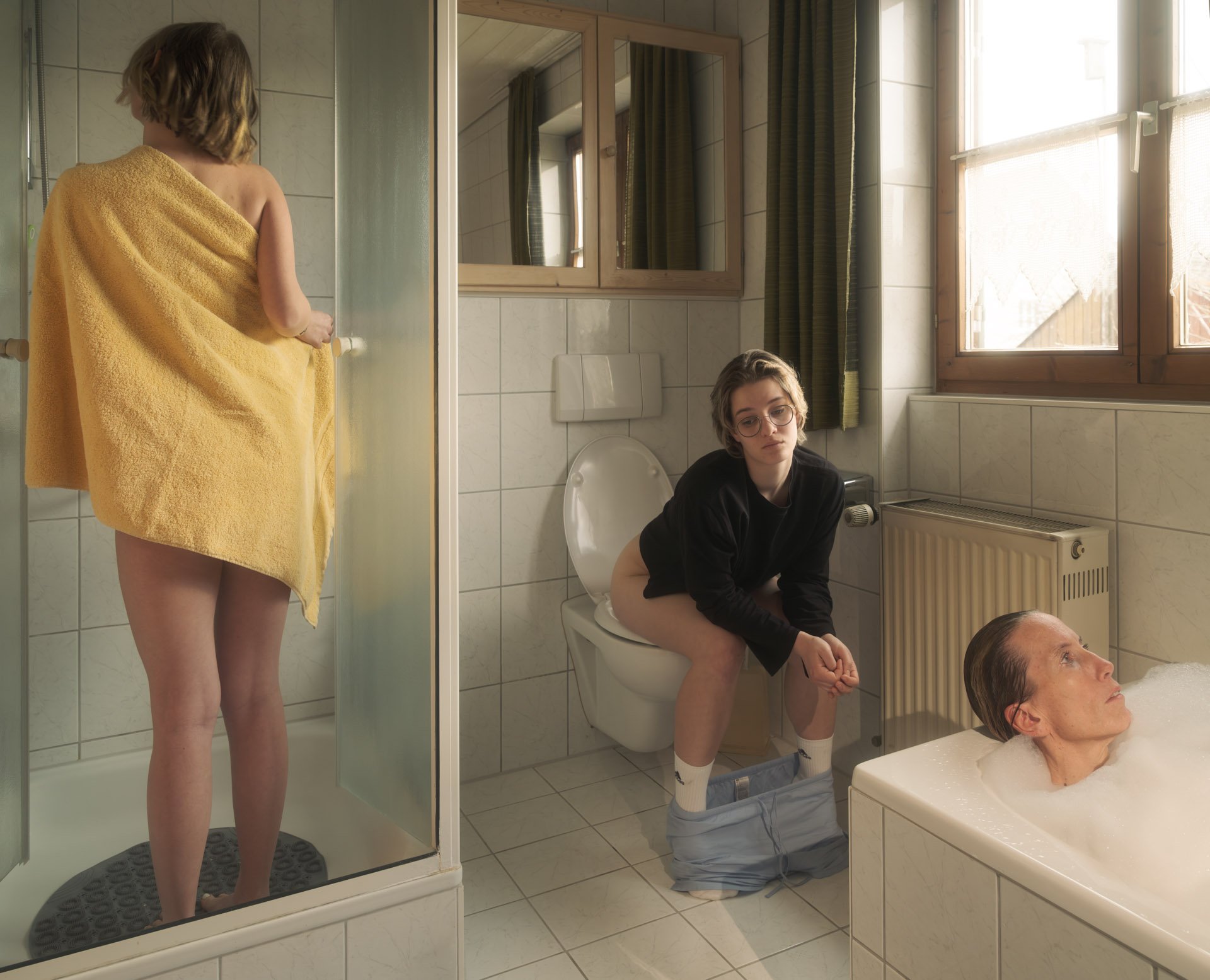 A woman in a yellow towel is standing in a shower, facing away. Inside the bathroom, a person is sitting on the toilet with pants down, looking at the person in the shower, with their head tilted back into a bathtub where another woman is lying with her head resting on the edge, hair wet, eyes closed, in a bubble bath. The bathroom has beige tiles, a window with curtains, and a mirror above the sink.