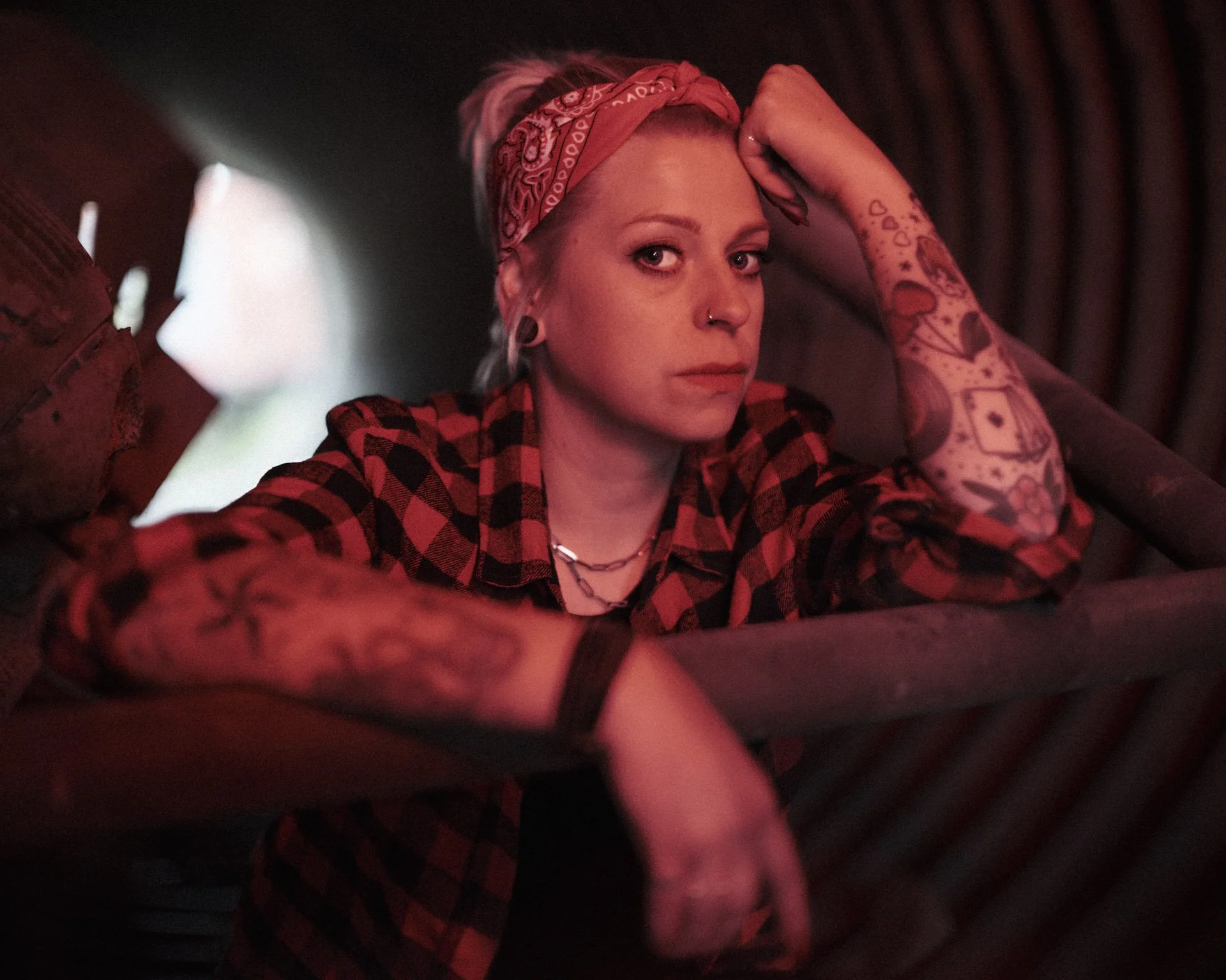 A woman with tattoos and a red bandana on her head, wearing a red and black plaid shirt, resting her arm on a motorcycle with a serious expression.