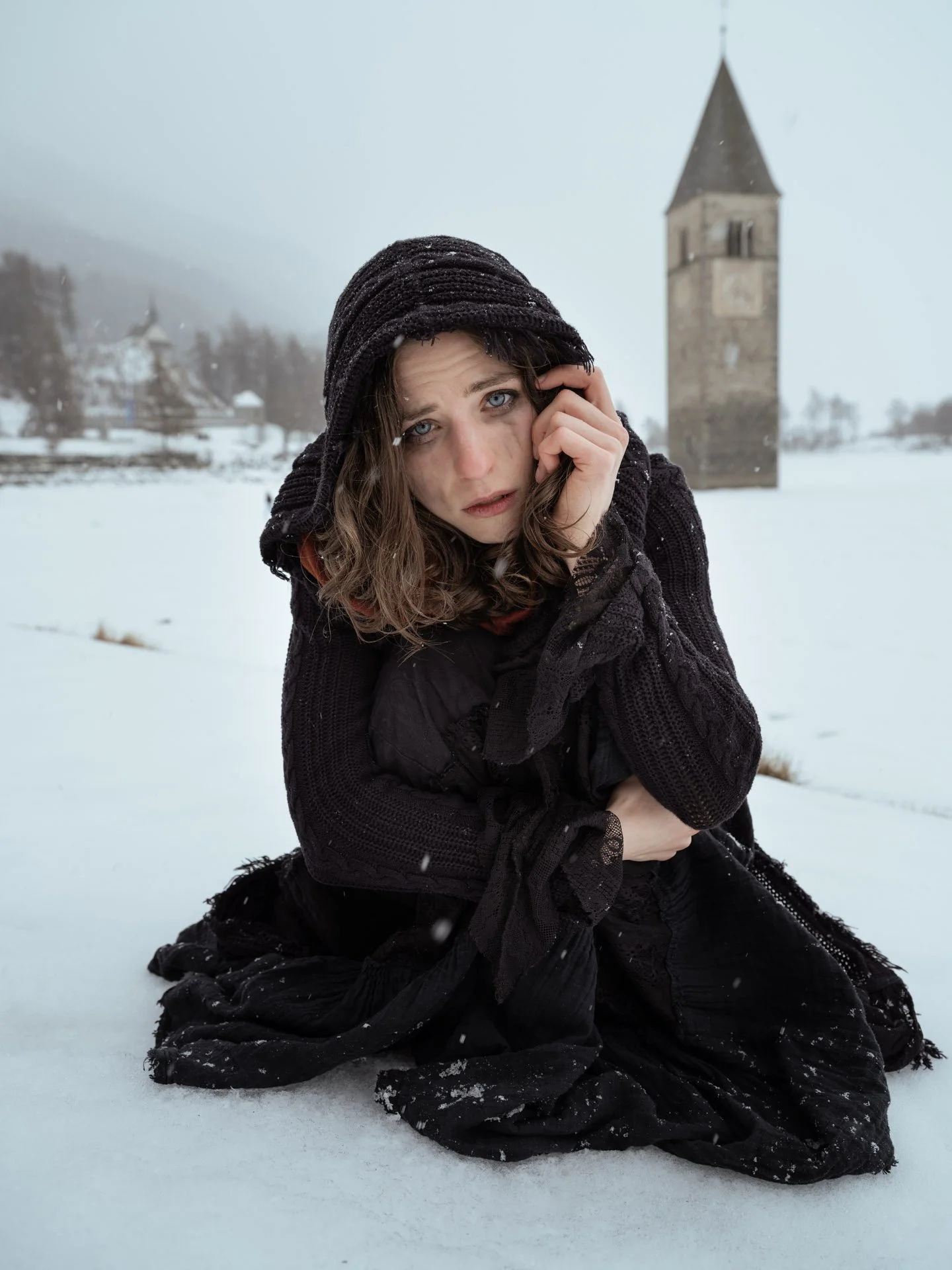 A woman with blue eyes and long brown hair, dressed in black, sitting in snow with a church steeple in the background, wearing a hood and looking at the camera.