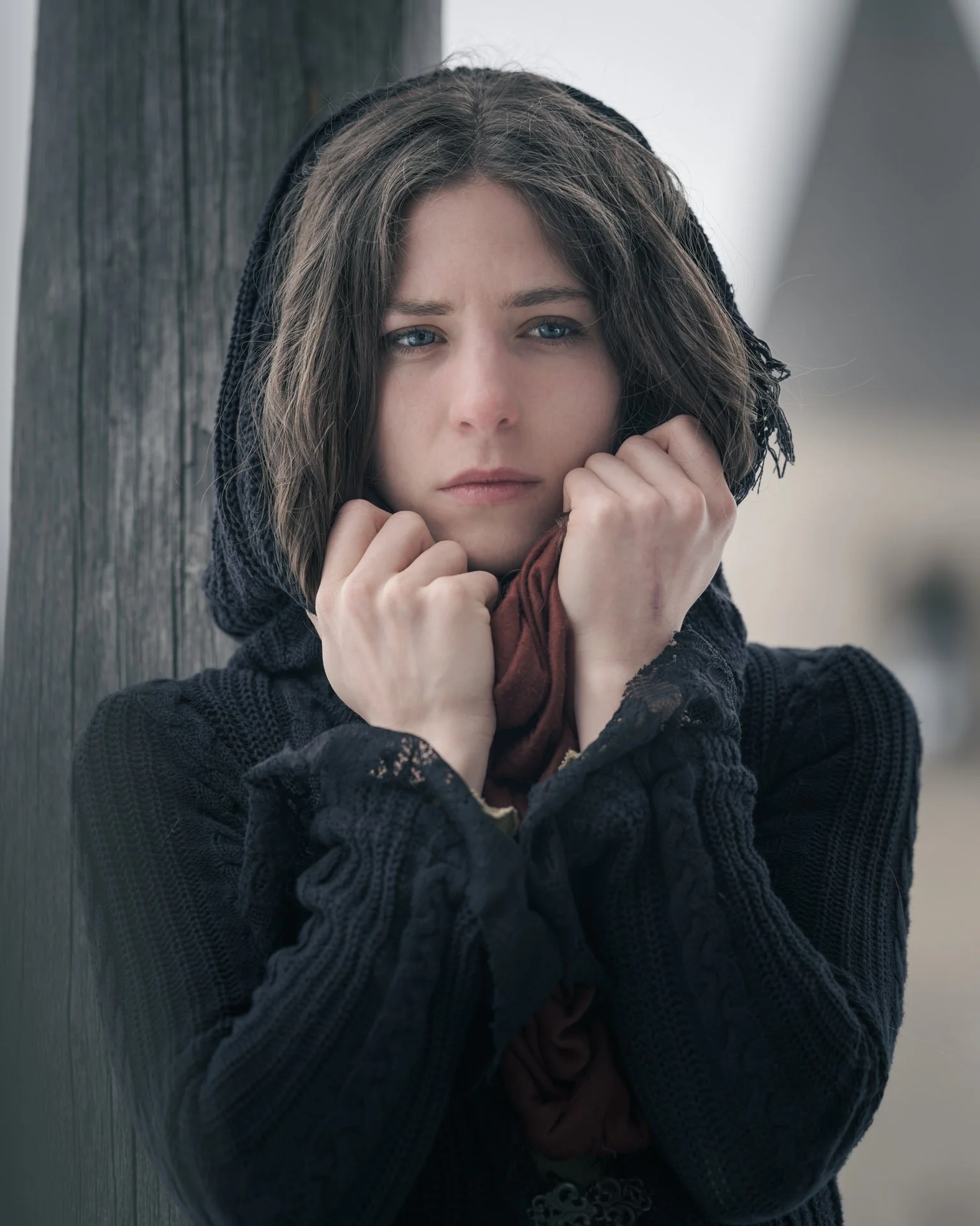 A woman with brown hair and blue eyes, wearing a black hoodie, is holding her hands close to her face, standing outdoors near a wooden post.