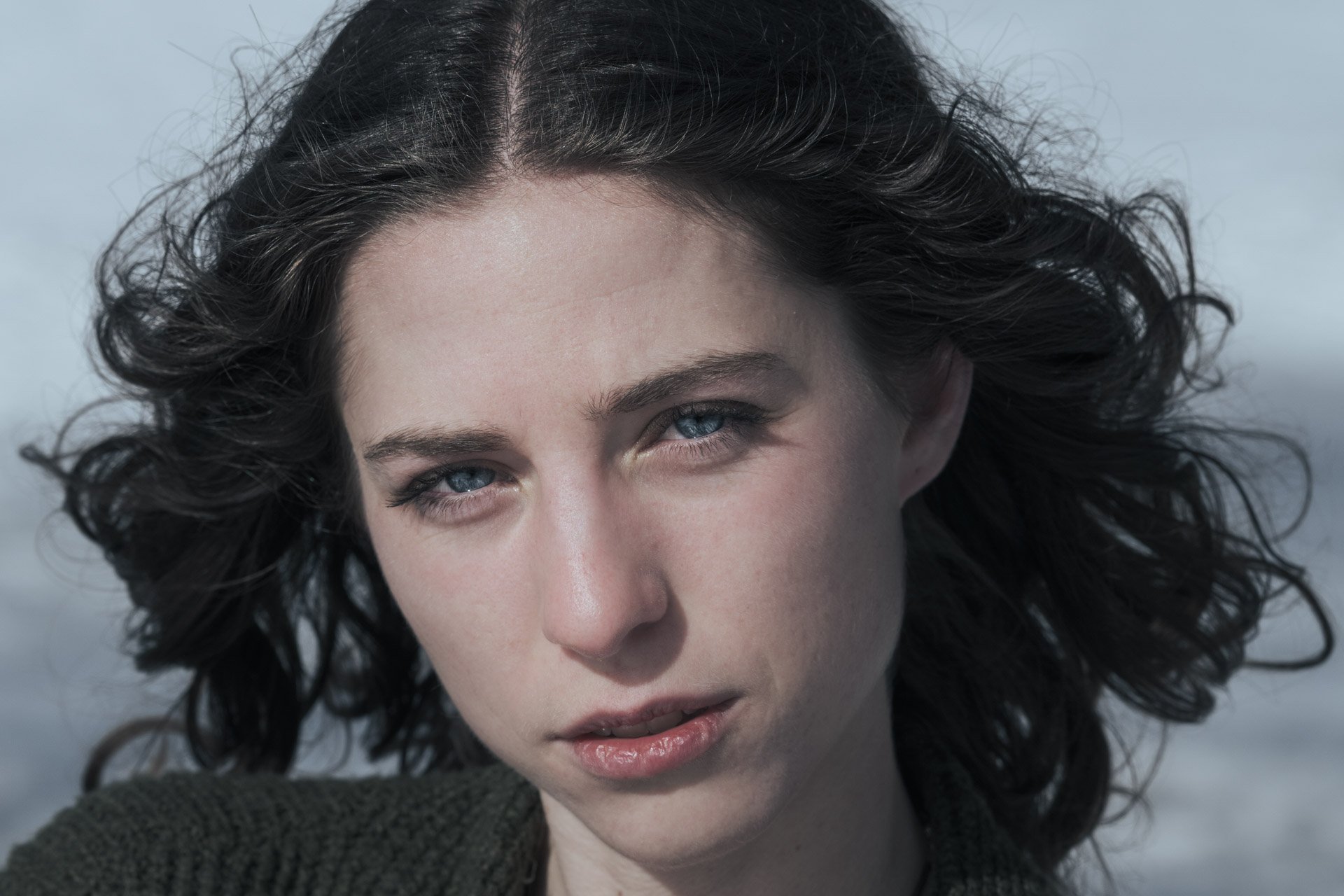 Close-up of a woman with curly dark hair and light skin, looking directly at the camera, with a neutral facial expression.