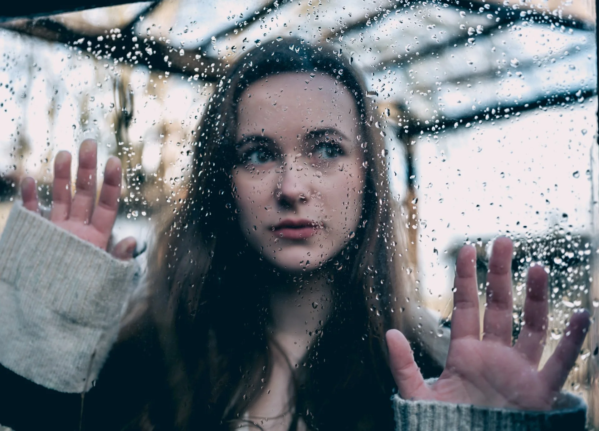 A young woman looking through a rain-covered window with droplets, touching the glass with both hands.