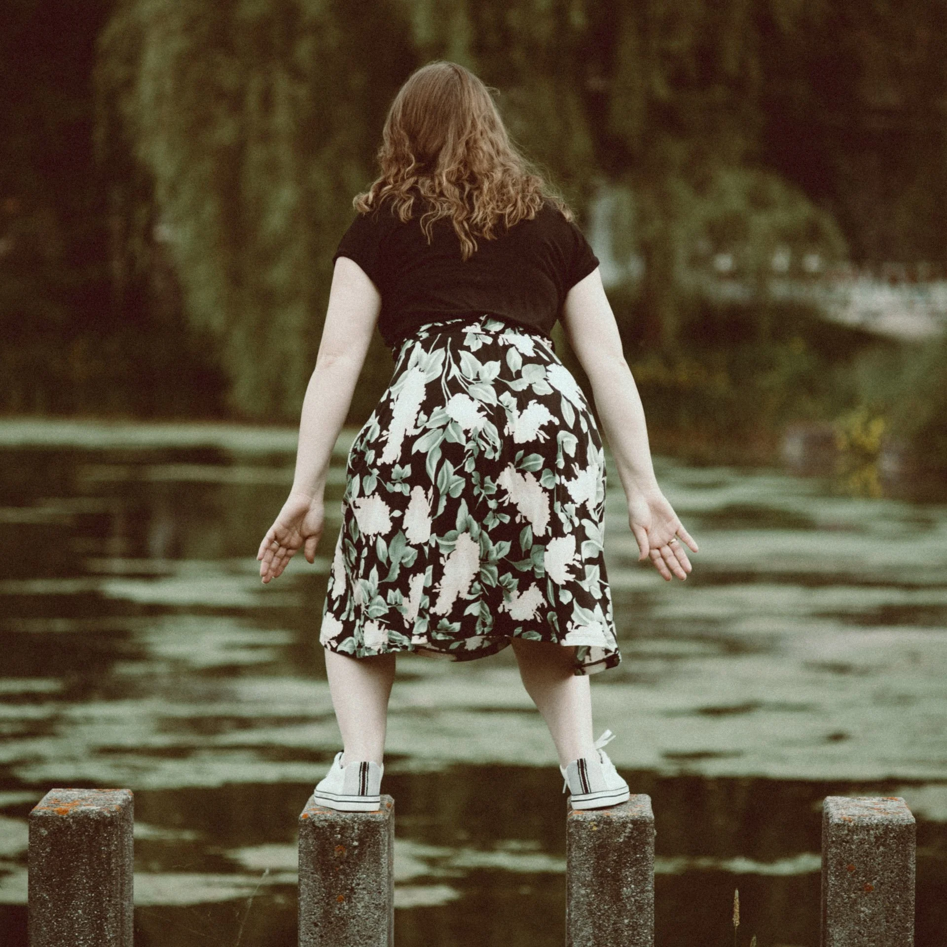 A woman with curly red hair standing on a wooden post near water, facing away from the camera, aiming to jump into the lake