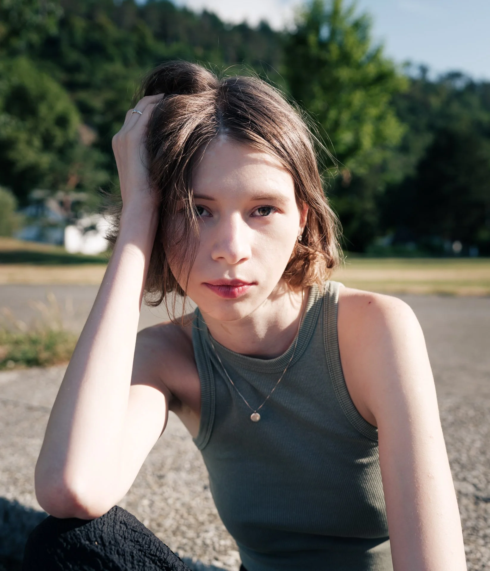 Close-up of a young woman with short, wavy brown hair and fair skin, wearing a gray tank top and a pearl necklace, sitting outdoors in a park with green trees and a clear blue sky in the background. She is touching her head with her left hand, lookin