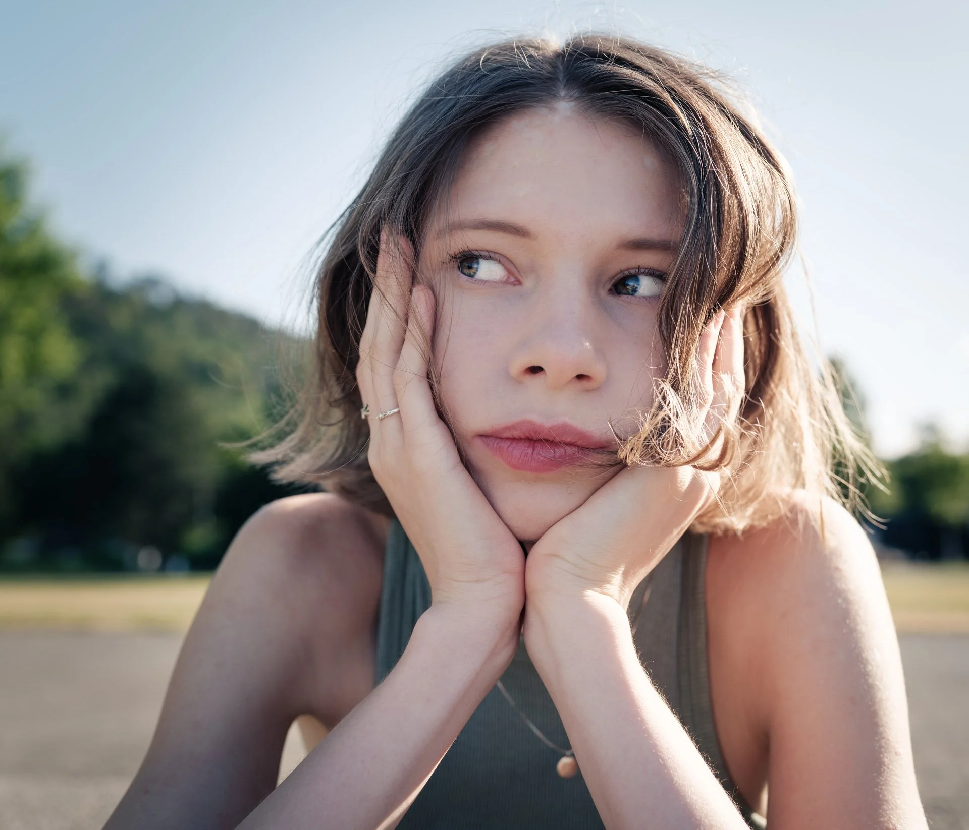 A young woman with light brown hair and blue eyes outdoors, holding her face with both hands, with a blurred background of trees and sky.
