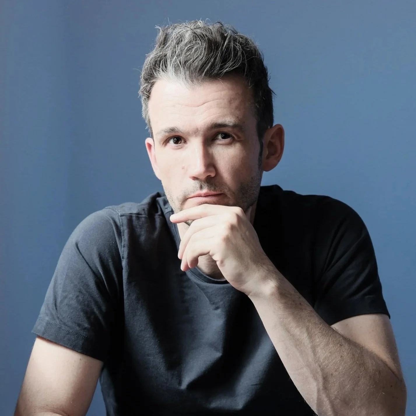 Portrait of a man with short, styled hair, wearing a black t-shirt, resting his chin on his hand, with a serious expression and a blue background.