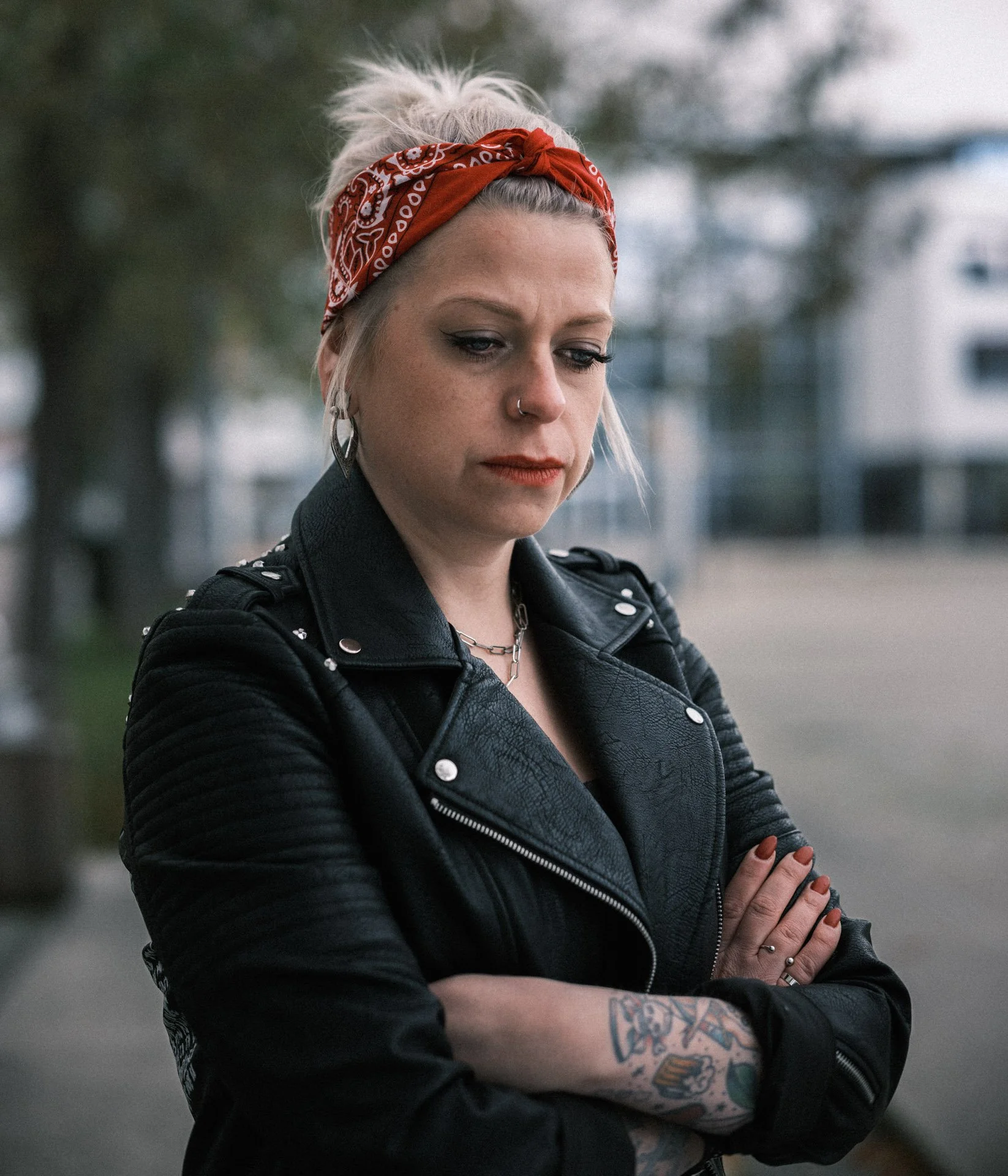 A young woman with platinum blonde hair wearing a red bandana, black leather jacket, multiple earrings, a nose ring, and a tattooed arm. She appears pensive with arms crossed outdoors.