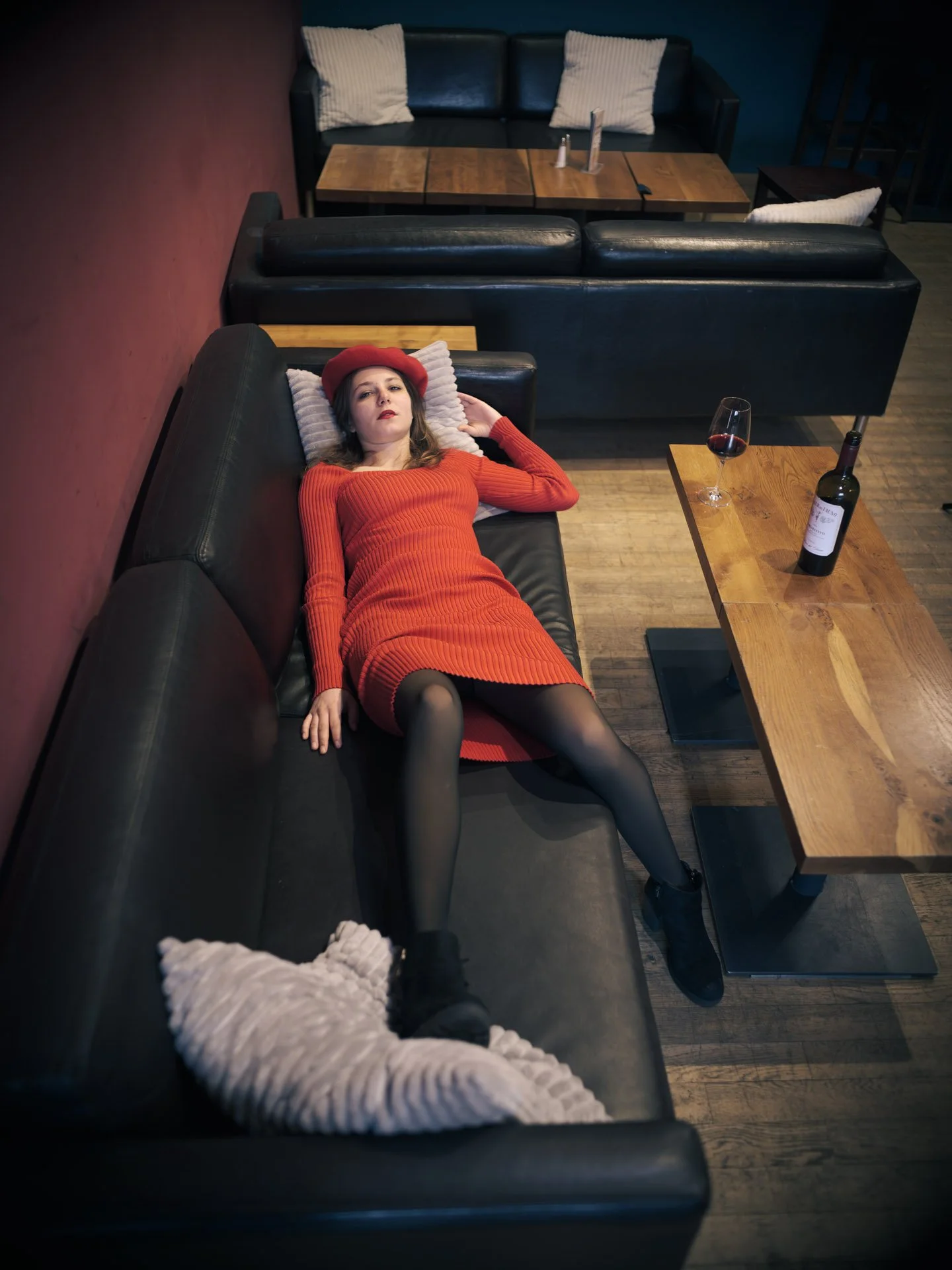 A woman dressed in red, wearing a red beret, lying on a black couch in a dimly lit room with wooden floors. She has a neutral expression and black stockings. A glass of red wine and a bottle of wine are on a wooden coffee table nearby. The room has a red and blue wall, and two additional couches with white pillows are visible.