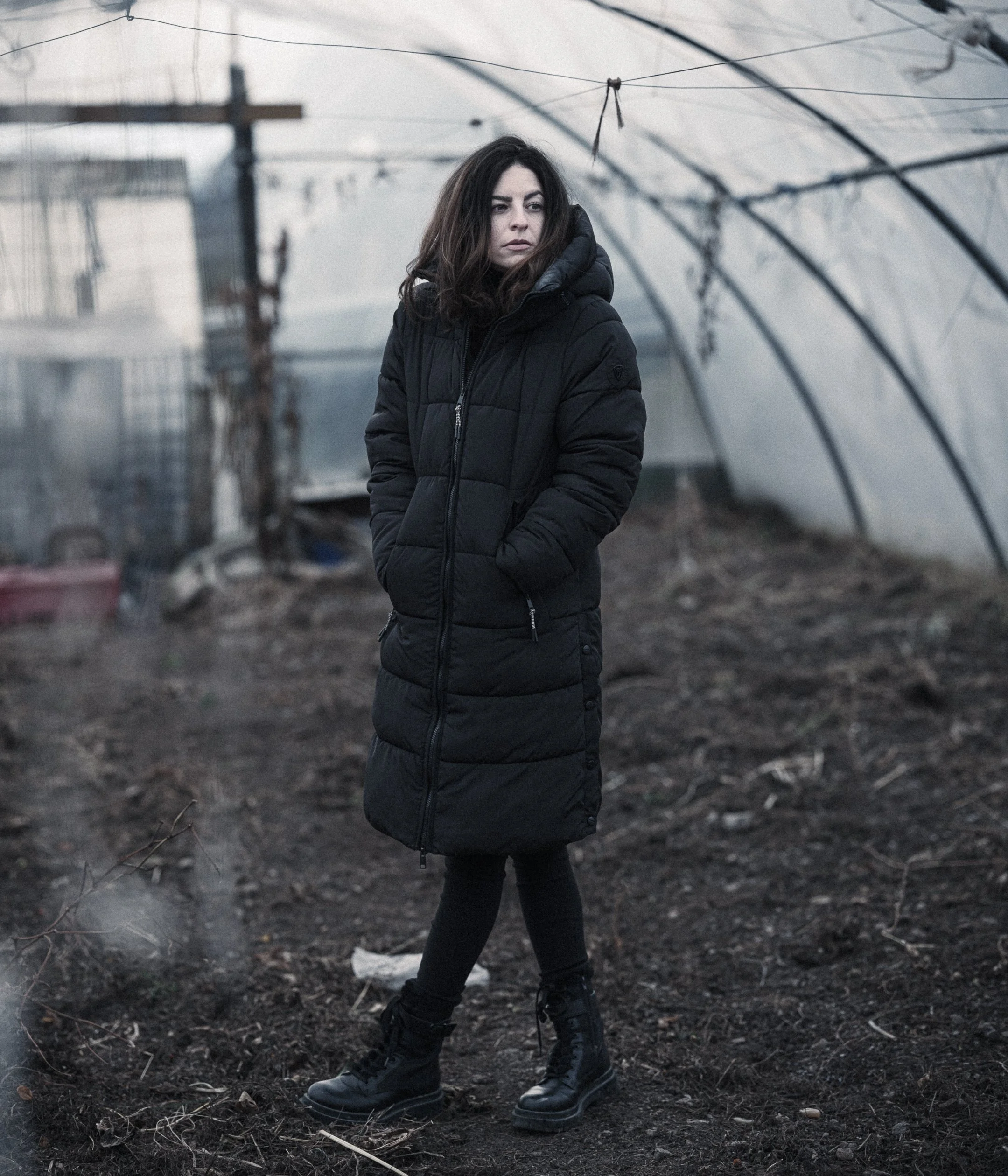 A woman in a black puffer jacket, black pants, and black boots stands outdoors on dark soil, with a plastic-covered greenhouse structure overhead and electric poles in the background, during overcast weather.