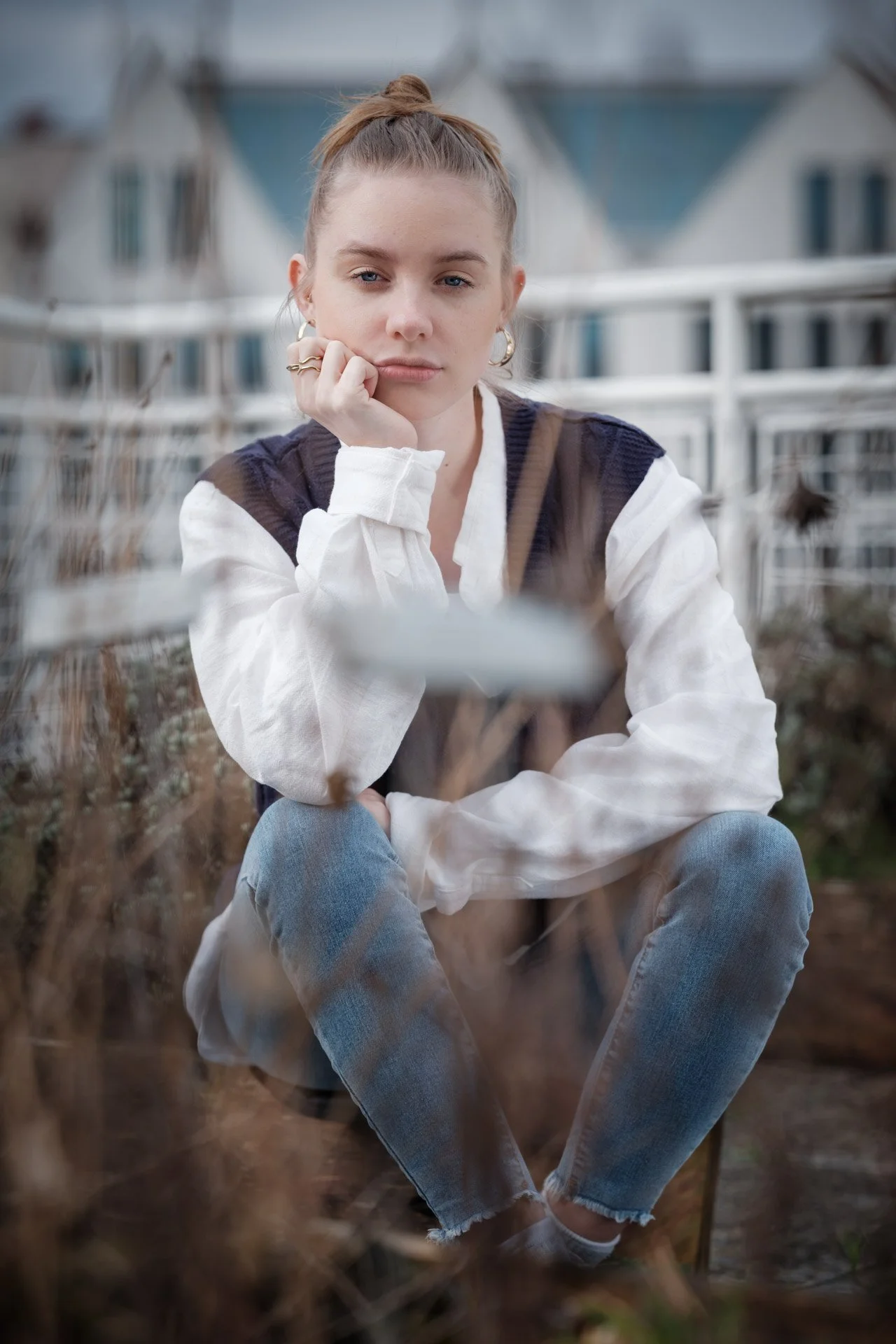 A young woman with brown hair in a bun, sitting outdoors among dried plants, wearing a white shirt with puffy sleeves, a dark vest, and light blue jeans, resting her chin on her hand and looking at the camera.