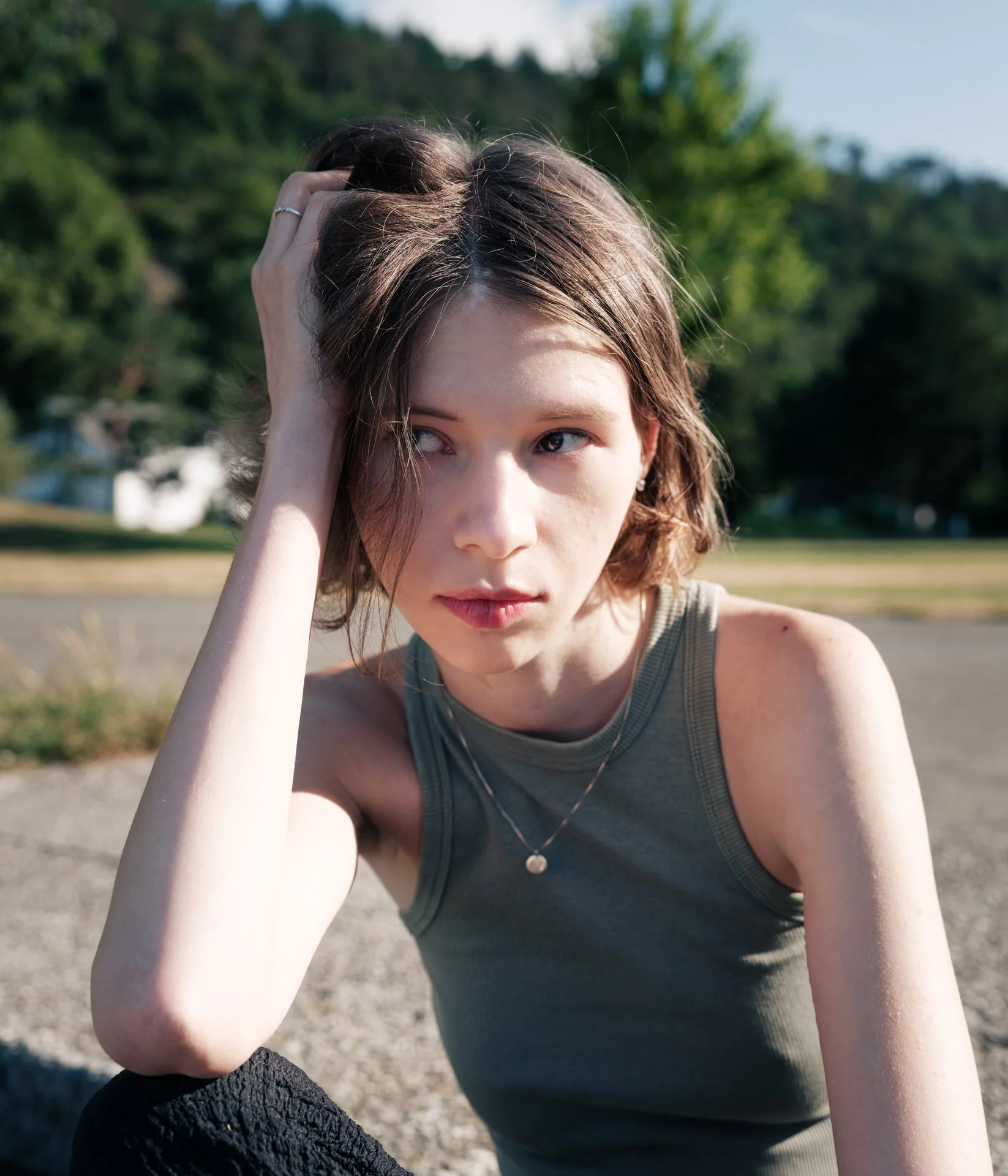 A young woman with light skin and brown hair sitting outdoors on a sunny day, touching her hair with her left hand, wearing a sleeveless top and a necklace, with trees and a grassy field in the background.