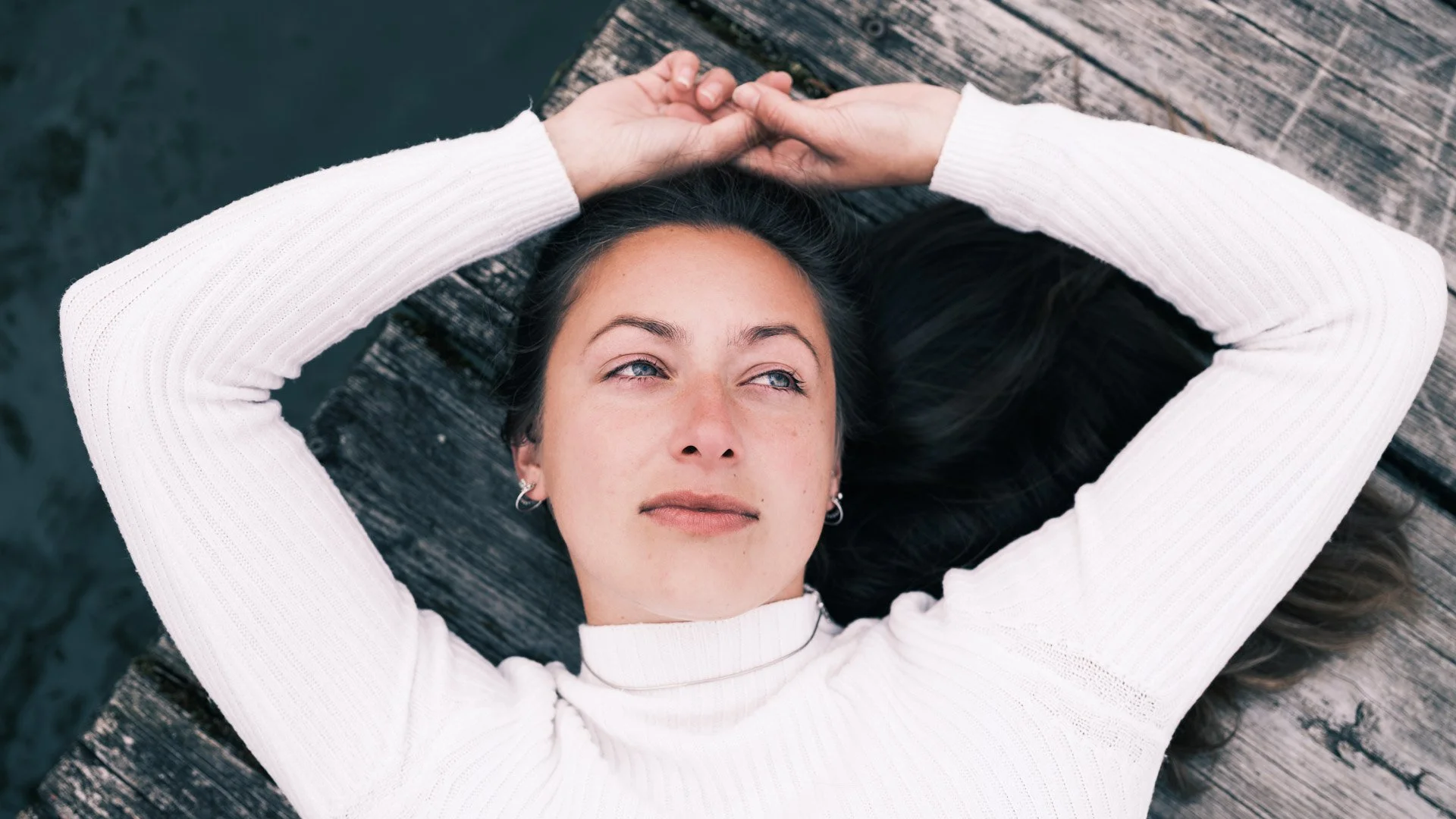 Woman lying on a wooden dock with her arms resting behind her head, looking up thoughtfully.