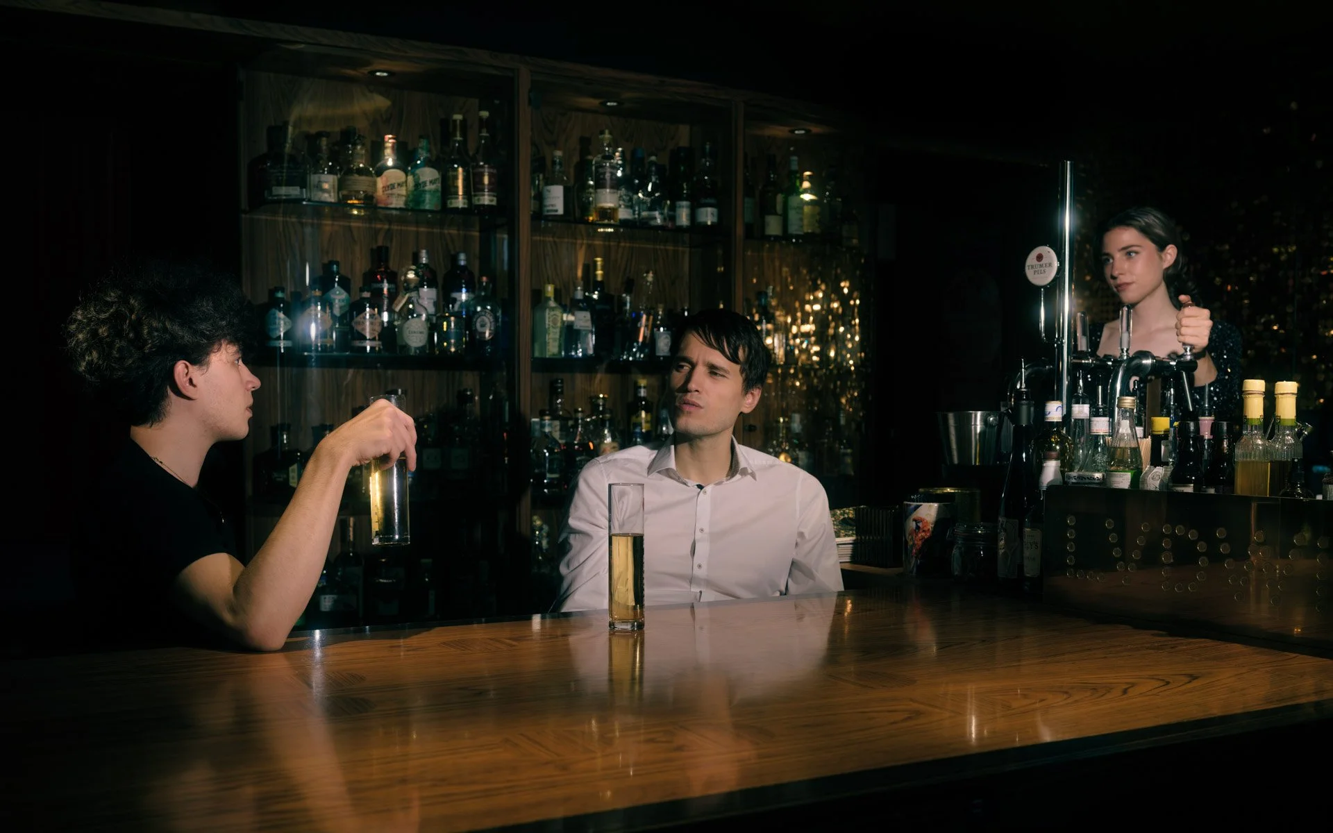 Two men sitting at a bar engaged in conversation, facing each other, with a female bartender in the background preparing a drink, behind a shelf of various liquor bottles, in a dimly lit setting.