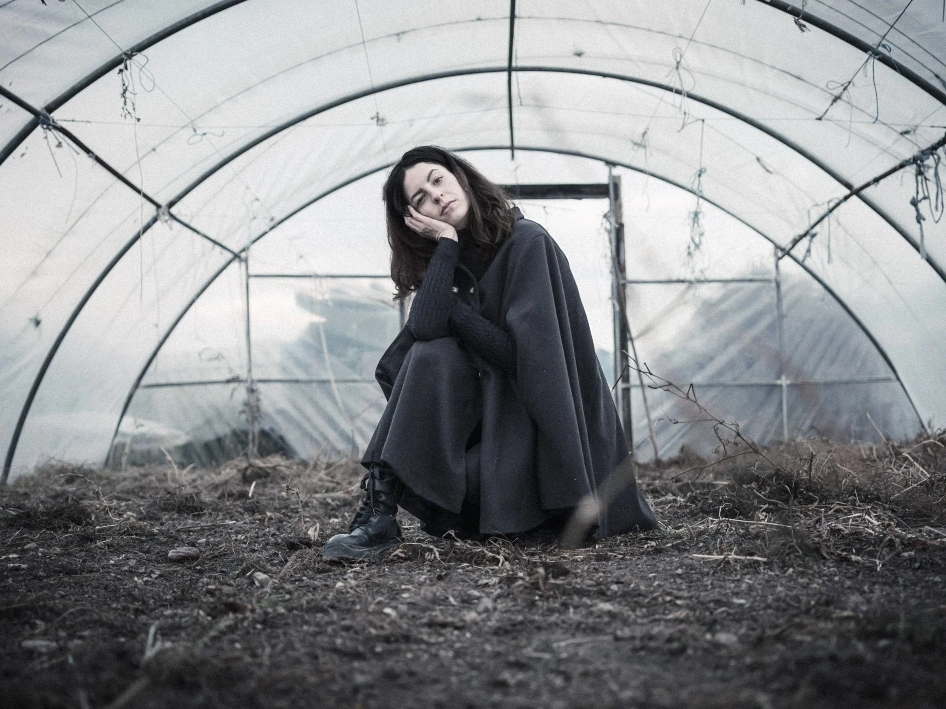 A young woman sitting inside a greenhouse with a dirt floor, looking contemplative with one hand resting on her face.