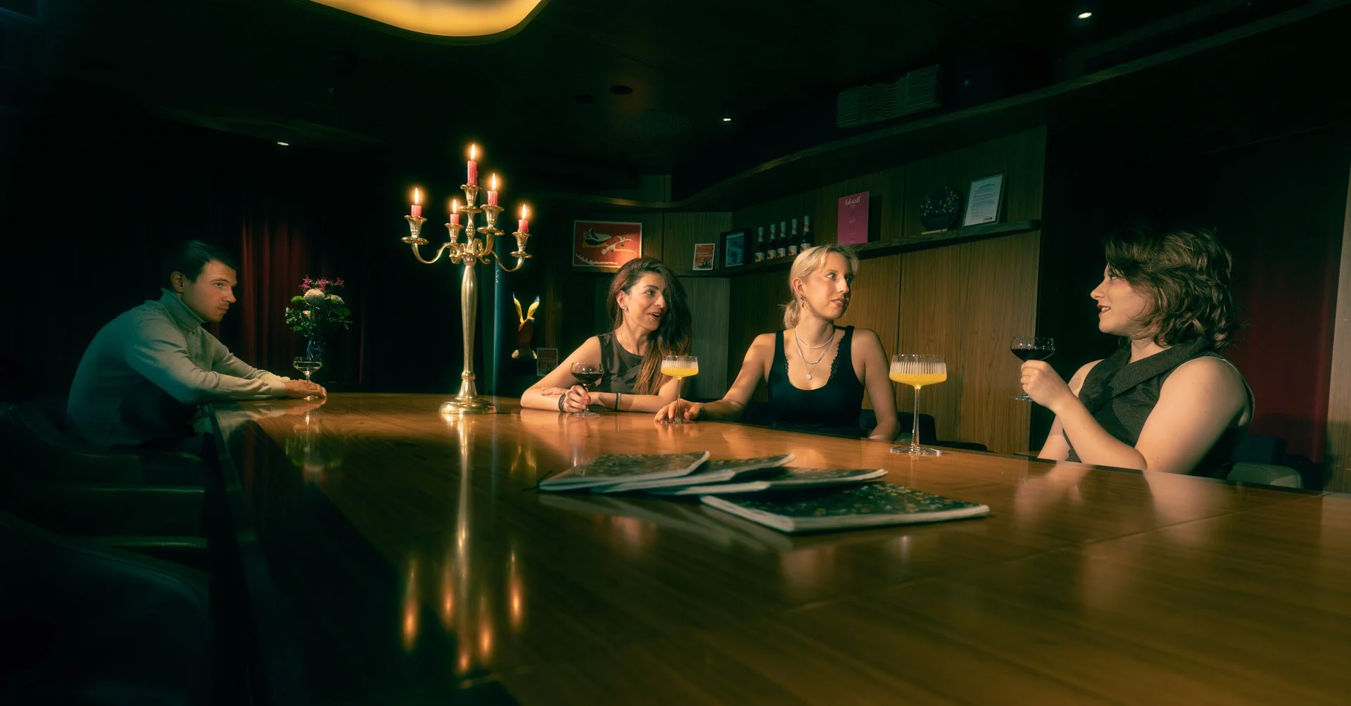 Four people sitting at a bar having drinks, with a man on the left and three women on the right, dark lighting, a chandelier, and magazines on the bar.