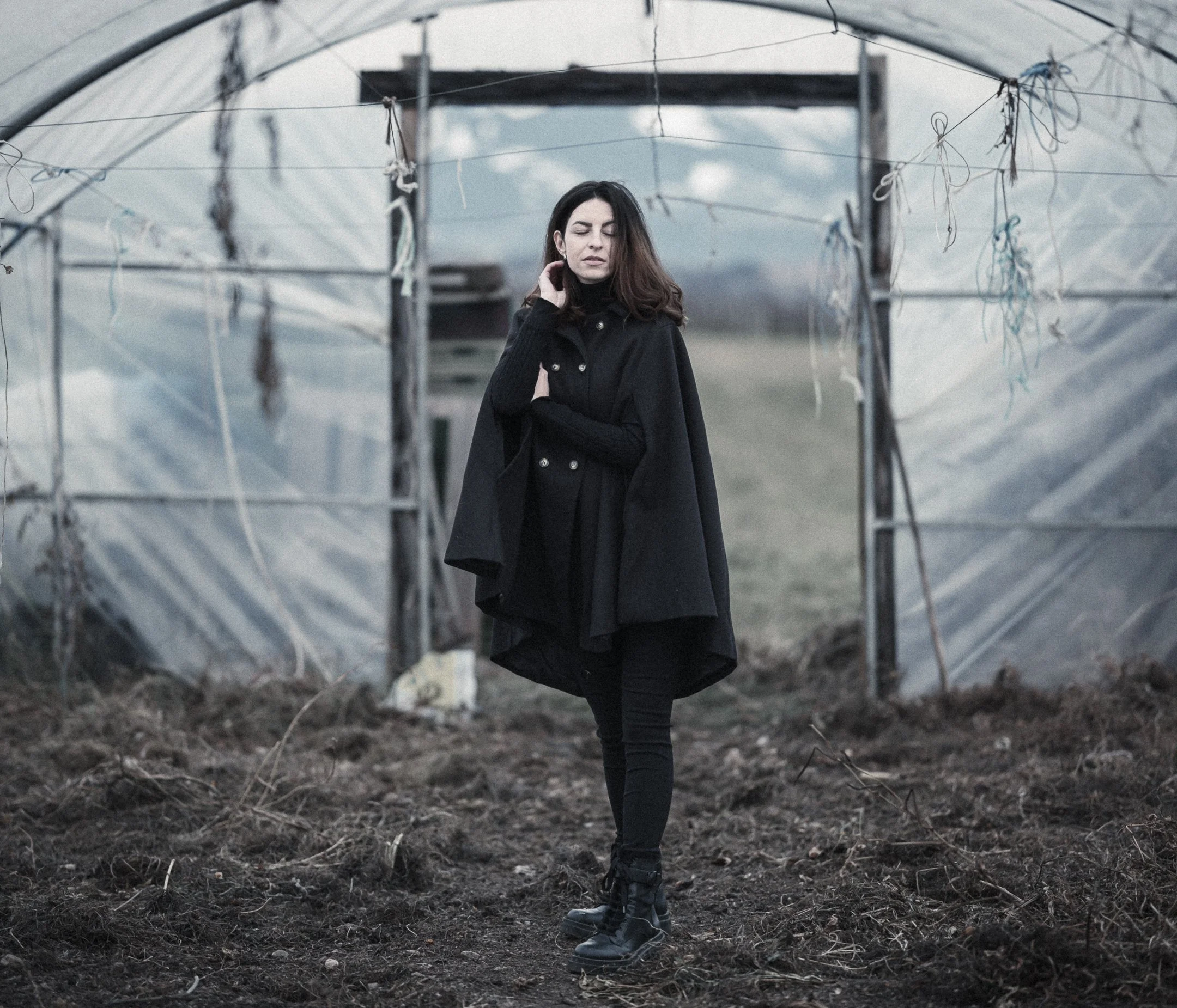 A woman with long dark hair, wearing a black coat and boots, standing in a greenhouse with a cloudy sky in the background.
