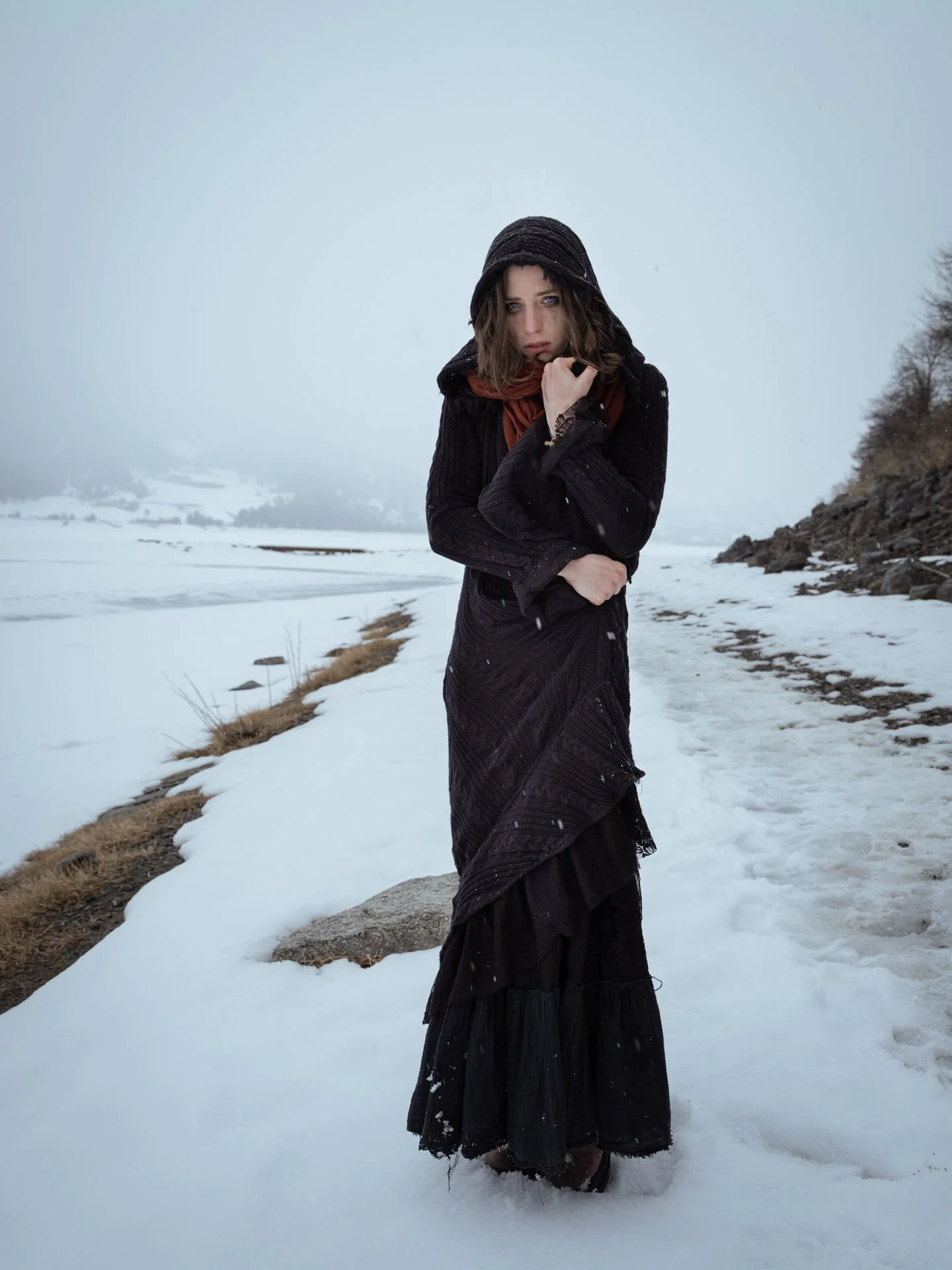 A woman standing in snow-covered landscape, dressed in black with a hood, holding her coat collar, with trees and a snow-covered field in the background.
