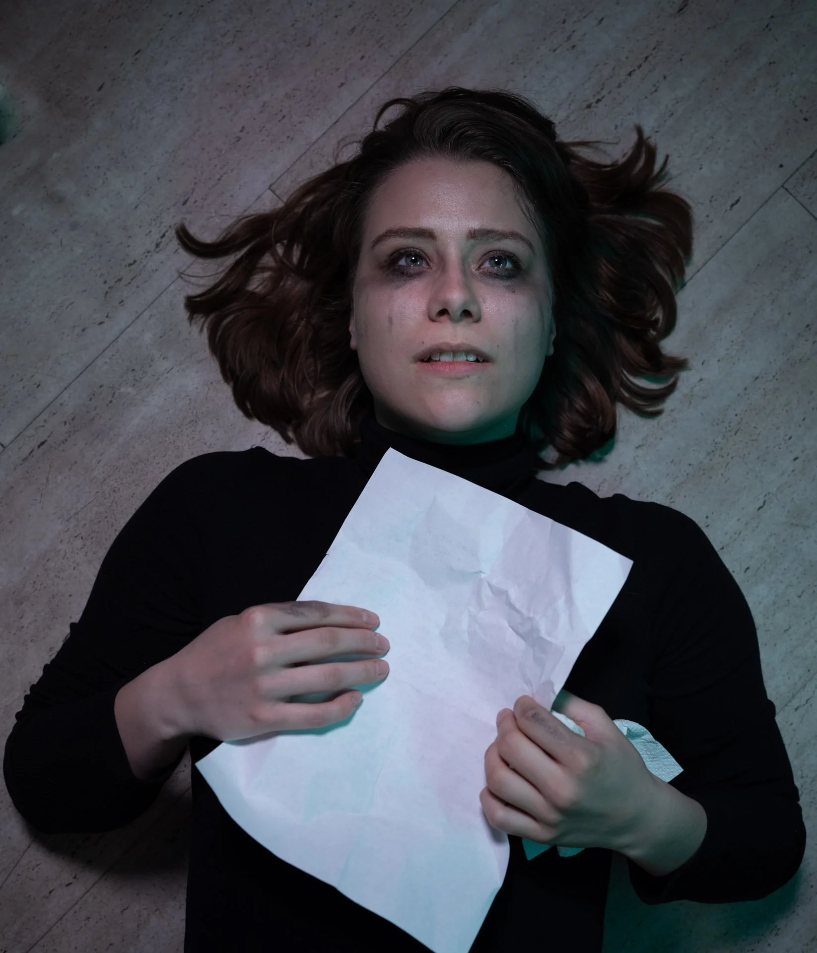 A woman with brown hair lying on a wooden floor, holding a crumpled piece of paper, with a distressed expression on her face.