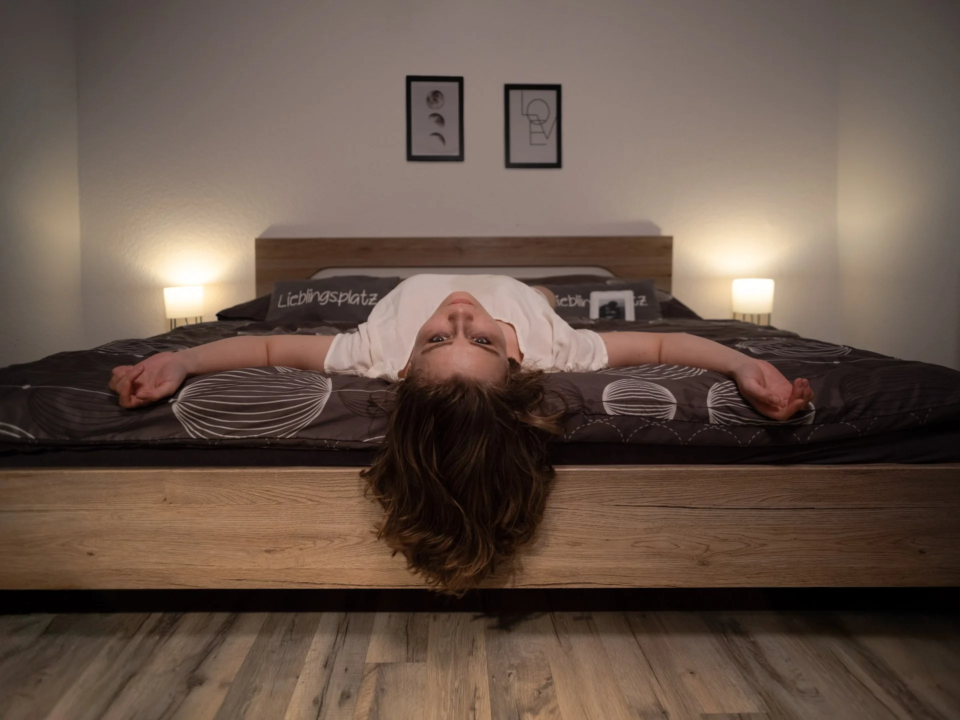 Young woman lying upside down on a bed with her head hanging over the edge, arms outstretched, in a bedroom with two lit lamps and framed artwork on the wall.