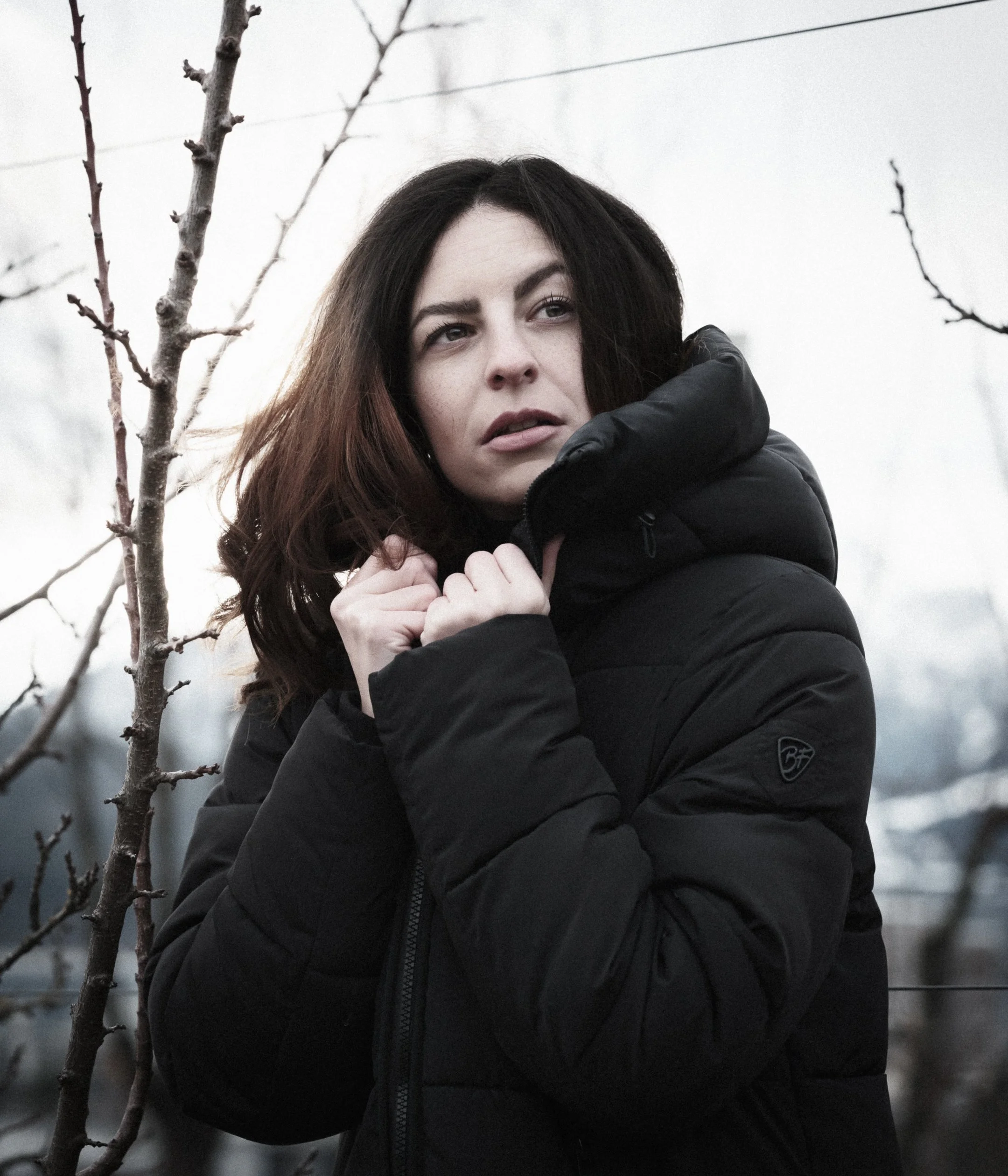 A woman with dark hair and fair skin wearing a black puffy jacket is outdoors, holding the collar of her jacket near her chin, with a background of leafless trees and gray sky.
